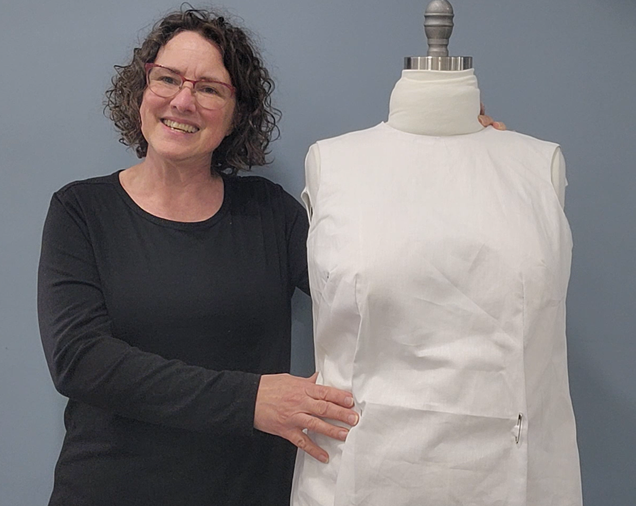 A woman with curly brown hair and glasses smiling, standing next to a white dress form or mannequin, against a plain gray background.