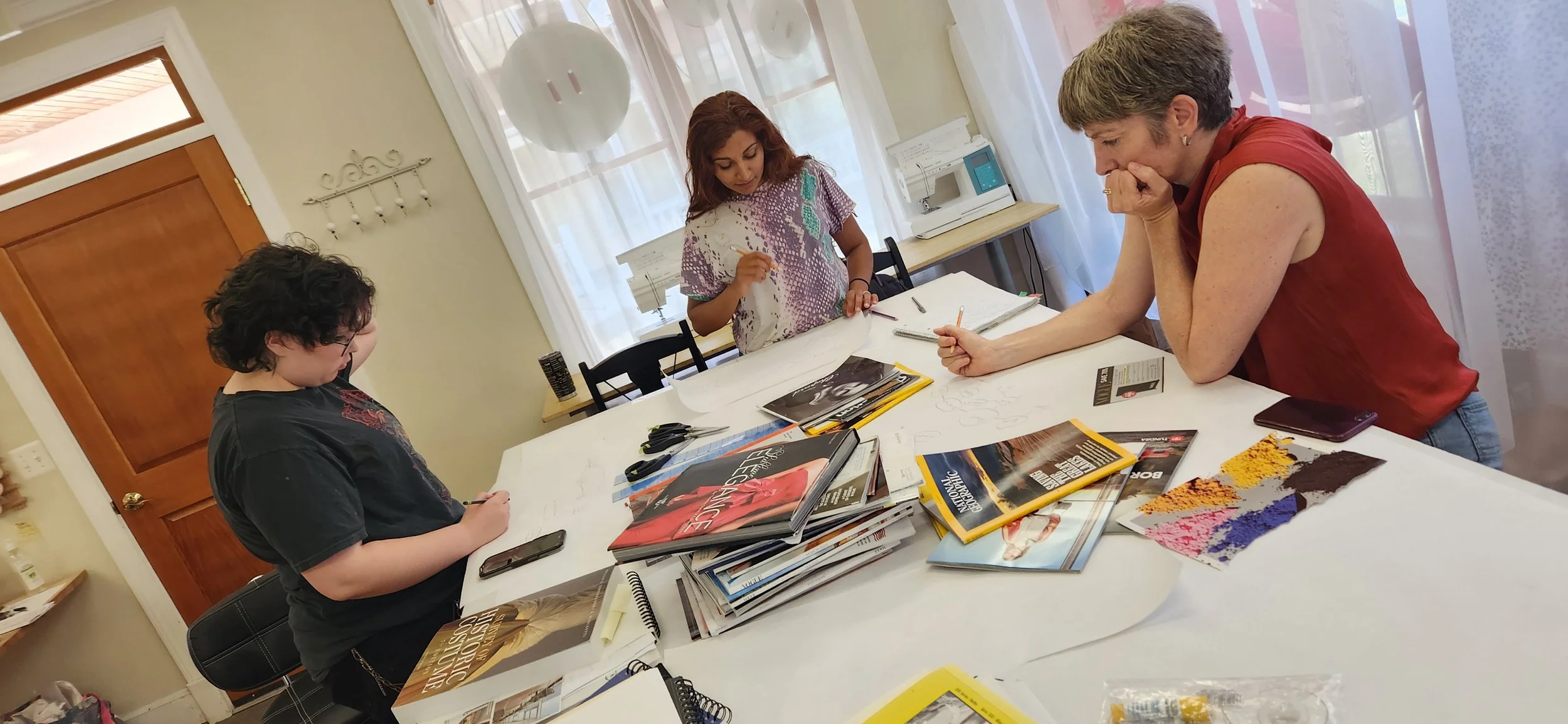 Three women around a table covered with magazines, papers, and art supplies, examining artwork and working on a creative project in a room with large windows and sheer curtains.