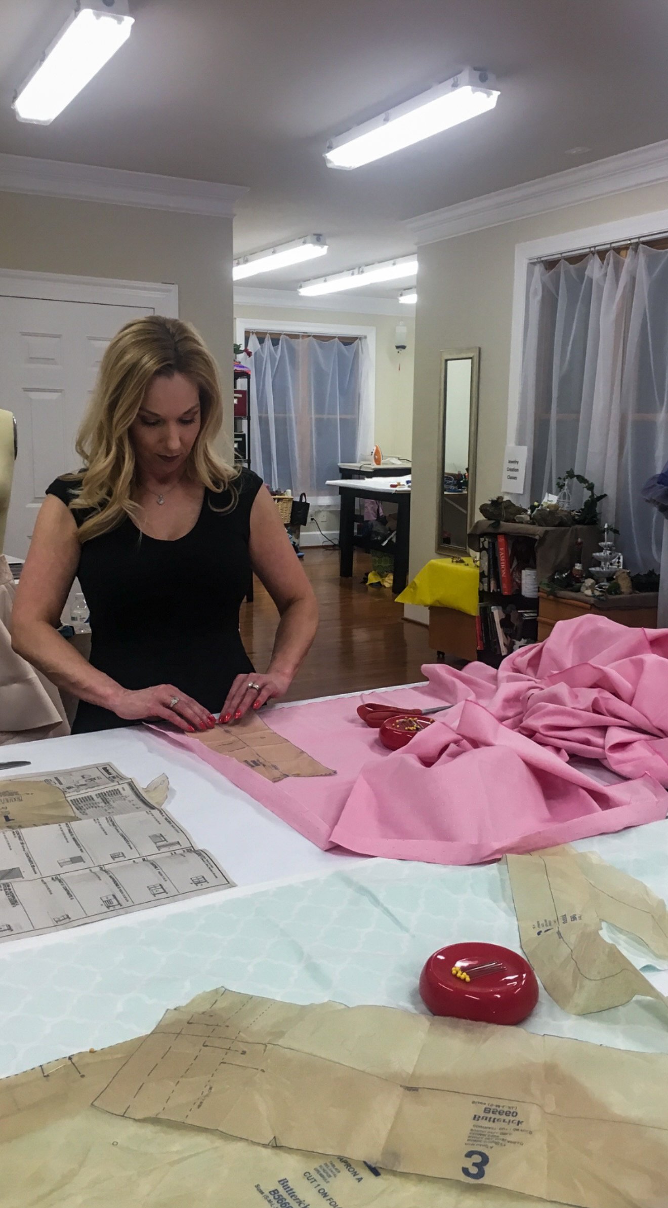 A woman working on a sewing project at a table with pink fabric and pattern paper in a well-lit room.