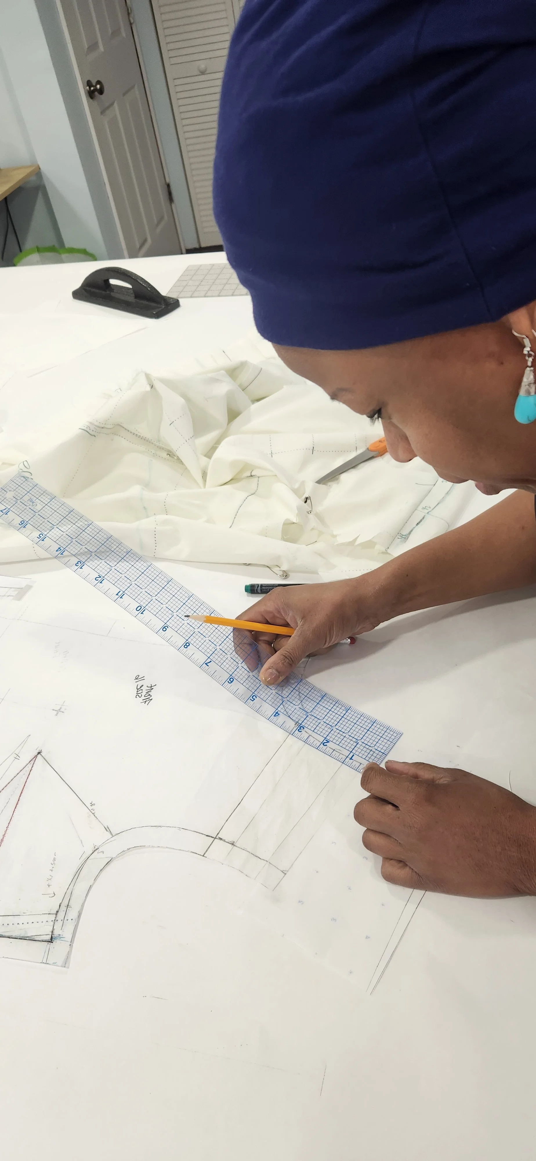 A person with a blue head covering and earrings, working on a large pattern or technical drawing on a table. They are using a ruler and pencil, with fabric and sewing tools nearby.