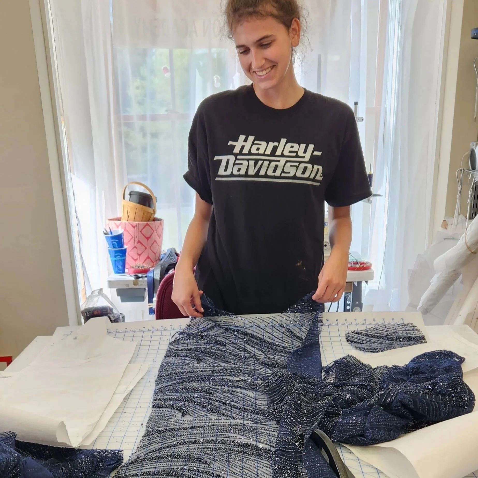 A young woman with curly hair wearing a black Harley-Davidson t-shirt is standing behind a table with various fabric pieces, a ruler, and papers, working on a sewing or craft project in a room with large windows and sheer curtains.