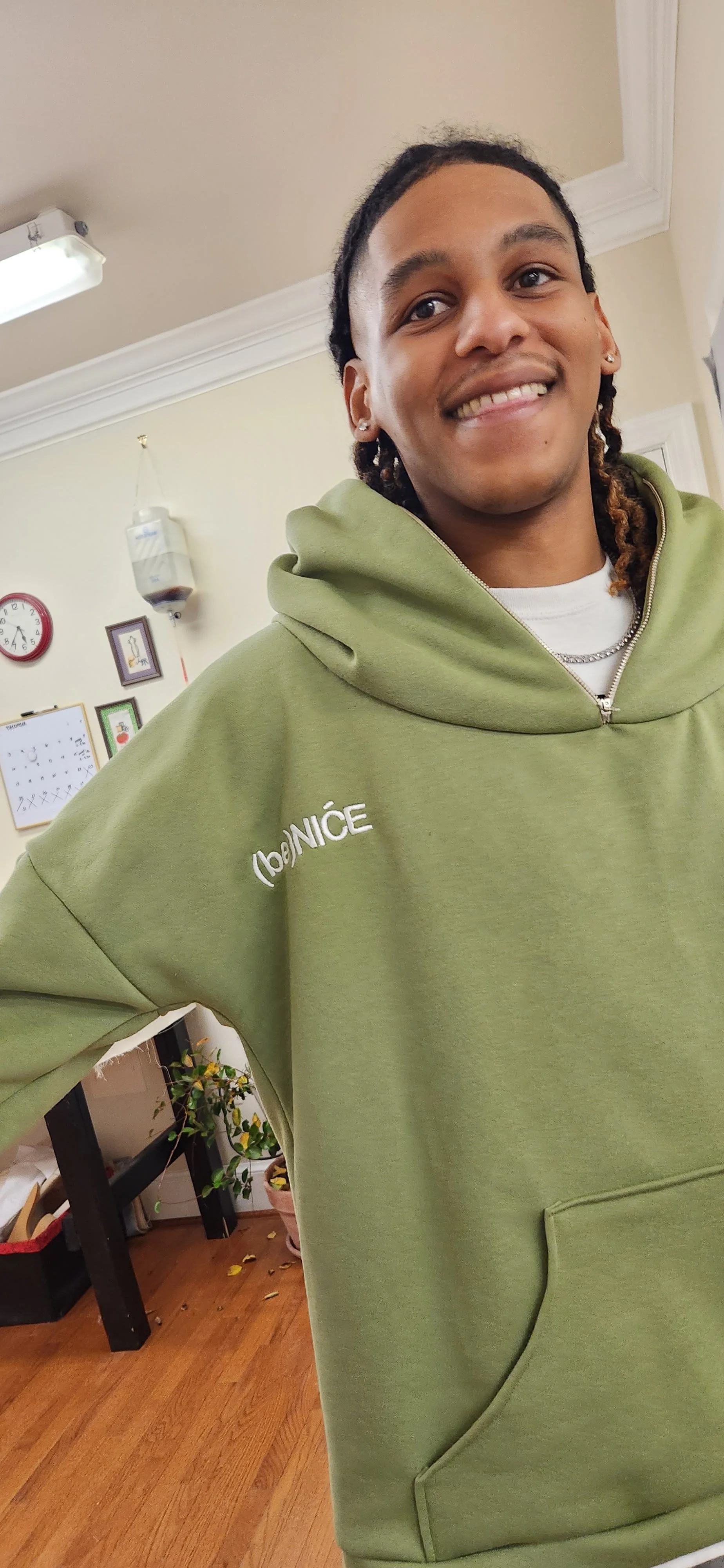 A young man with dreadlocks smiling, wearing a green hoodie with the word 'beNICE' on it, inside a room with a wall clock, framed pictures, and potted plants in the background.