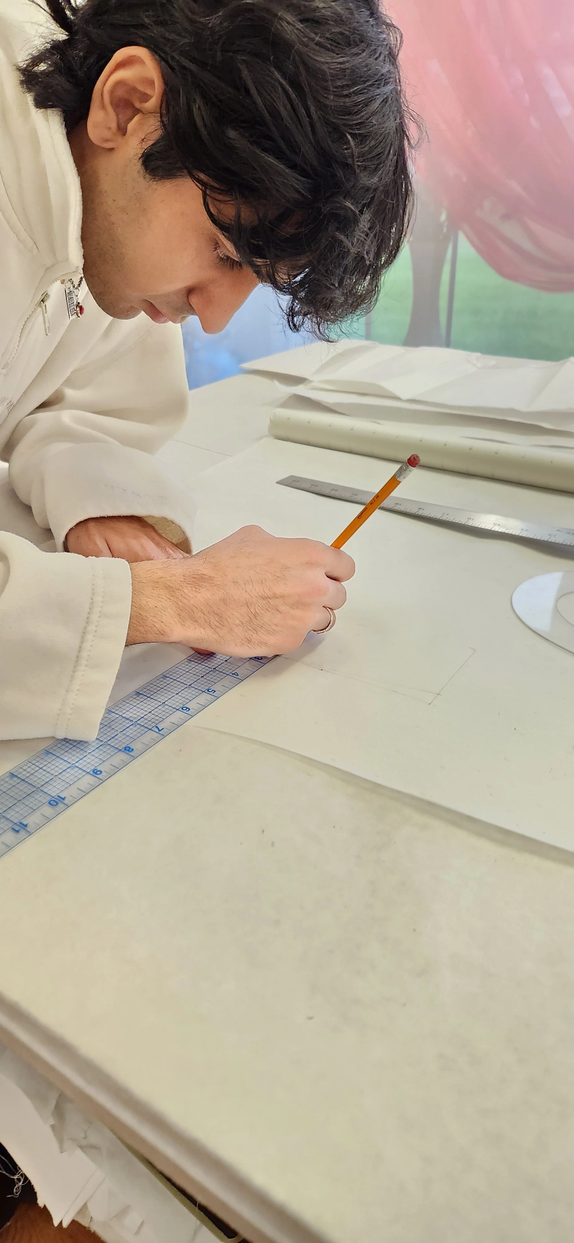 A person with curly dark hair wearing a cream hoodie is drawing on a piece of paper with a pencil and ruler on a white table.