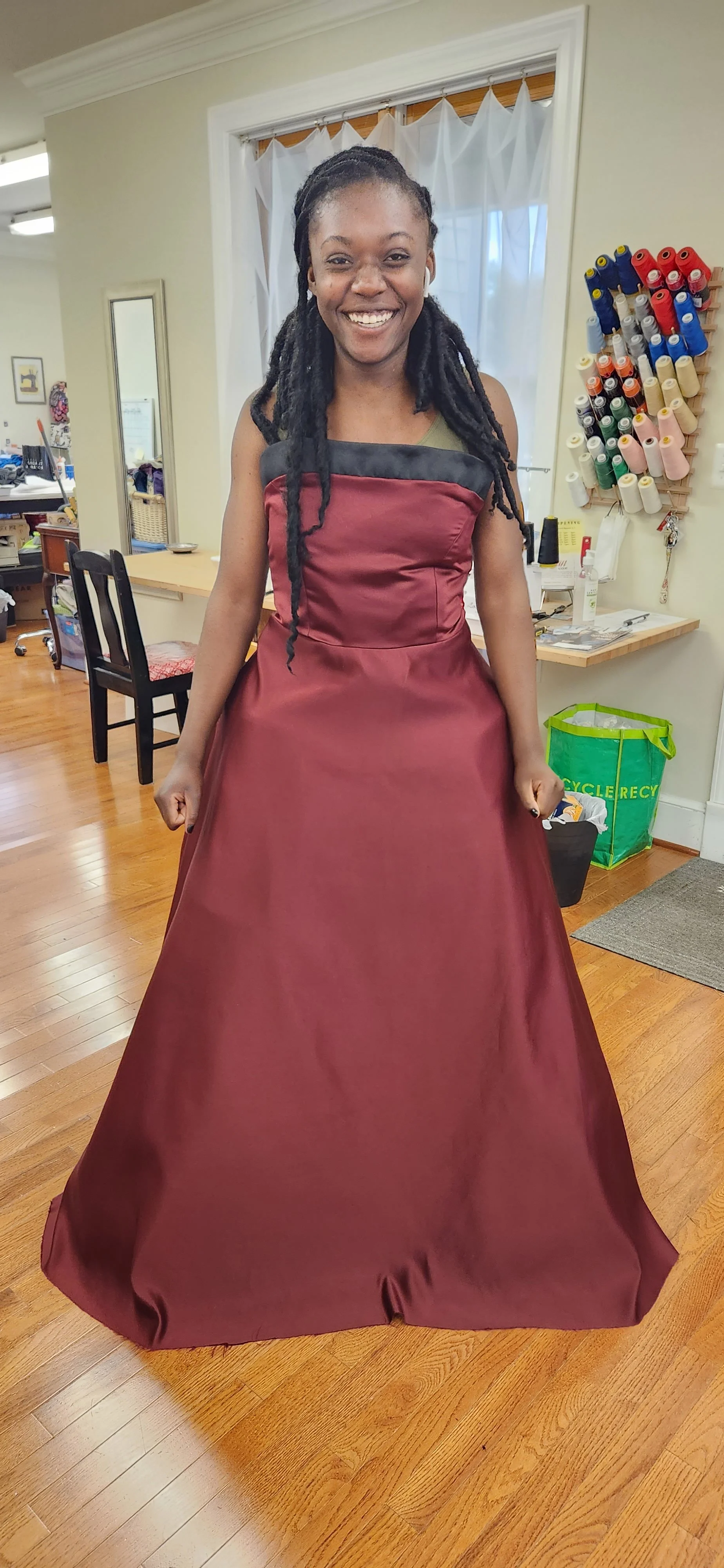 Woman smiling in a maroon gown inside a room with a hardwood floor, sewing supplies, and a green recycling bag.