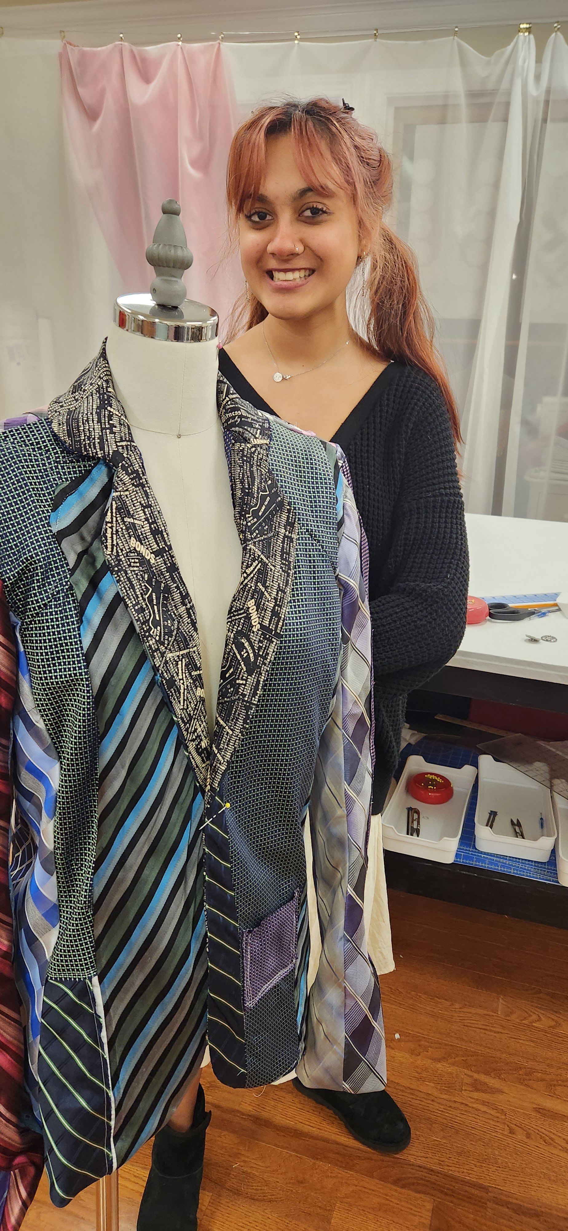 A woman smiling in a workspace, standing next to a mannequin dressed with various fabric swatches and patterns, with sewing tools and supplies on the table behind her.
