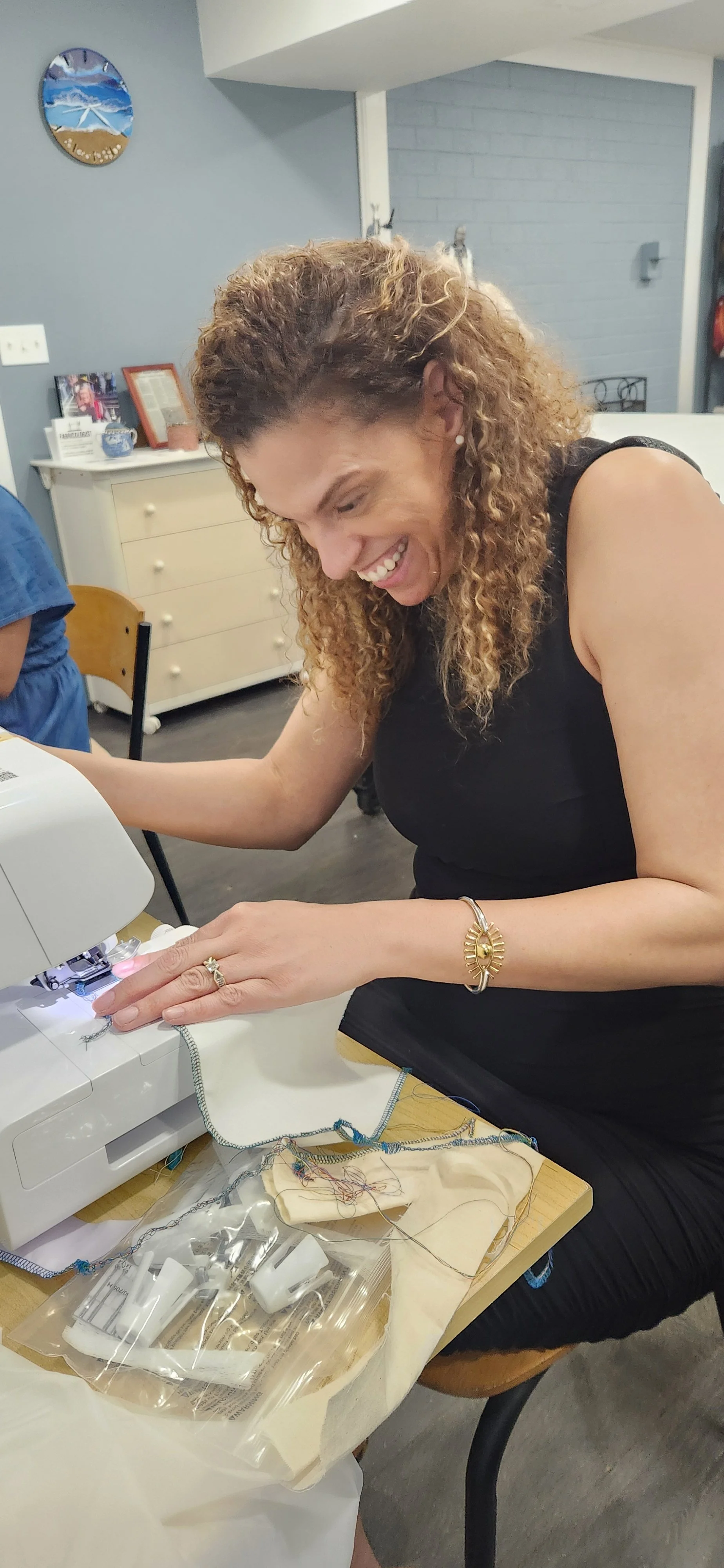 Woman sewing fabric on a machine, smiling and looking down, with sewing supplies and fabric on a table.