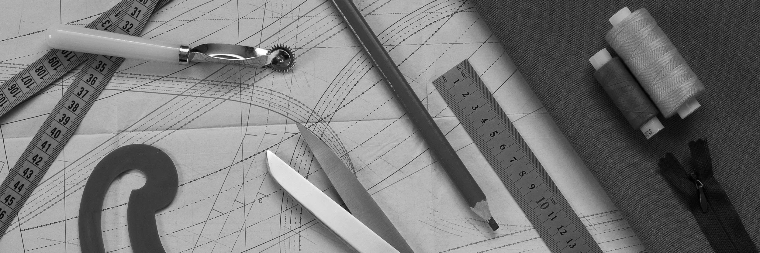 Drawing tools including a protractor, a ruler, a pencil, a curved ruler, a utility knife, and sewing spools on a technical sketch or pattern.