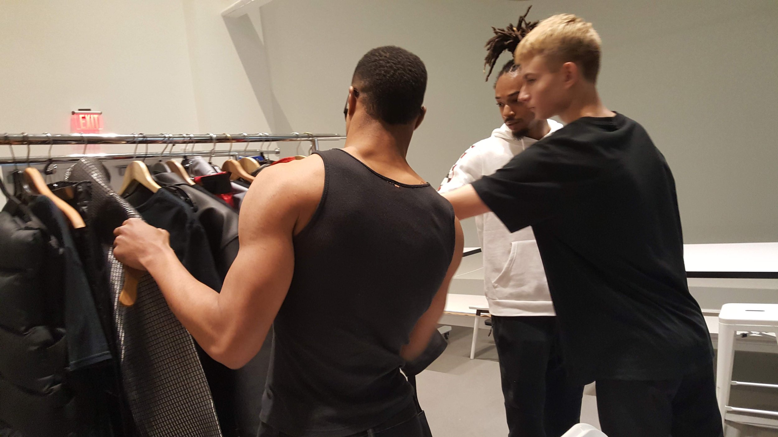 Three young men browse a clothing rack in a store or dressing room, with one picking up a black shirt, and the other two looking at the clothing.