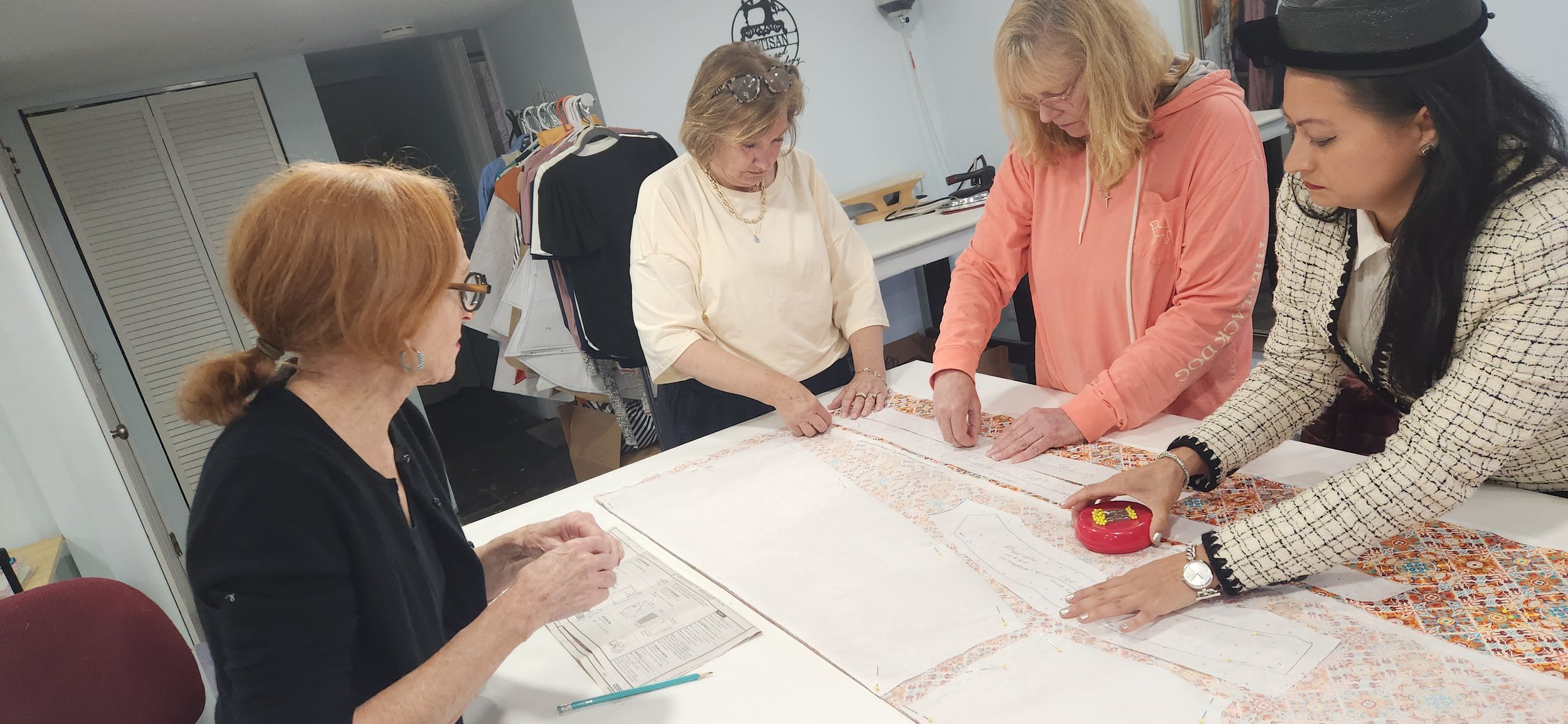 Four women gathered around a table, reviewing fabric and patterns for sewing or fashion design in a workroom.