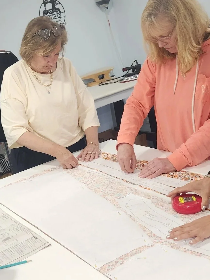 Two women are working together on a fabric project, measuring and pinning fabric on a table. One woman is wearing a cream shirt and sunglasses on her head; the other is in a peach jacket. Various sewing tools are on the table.