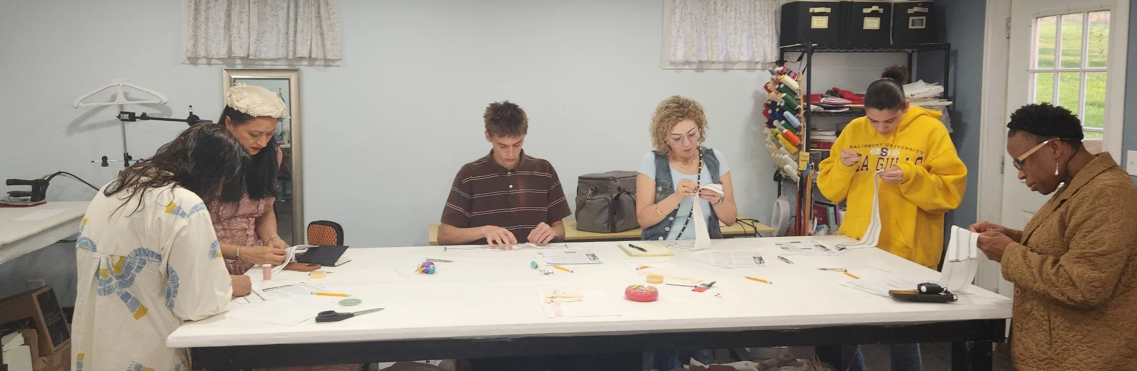 Group of six people working on sewing projects around a large table in a craft room.