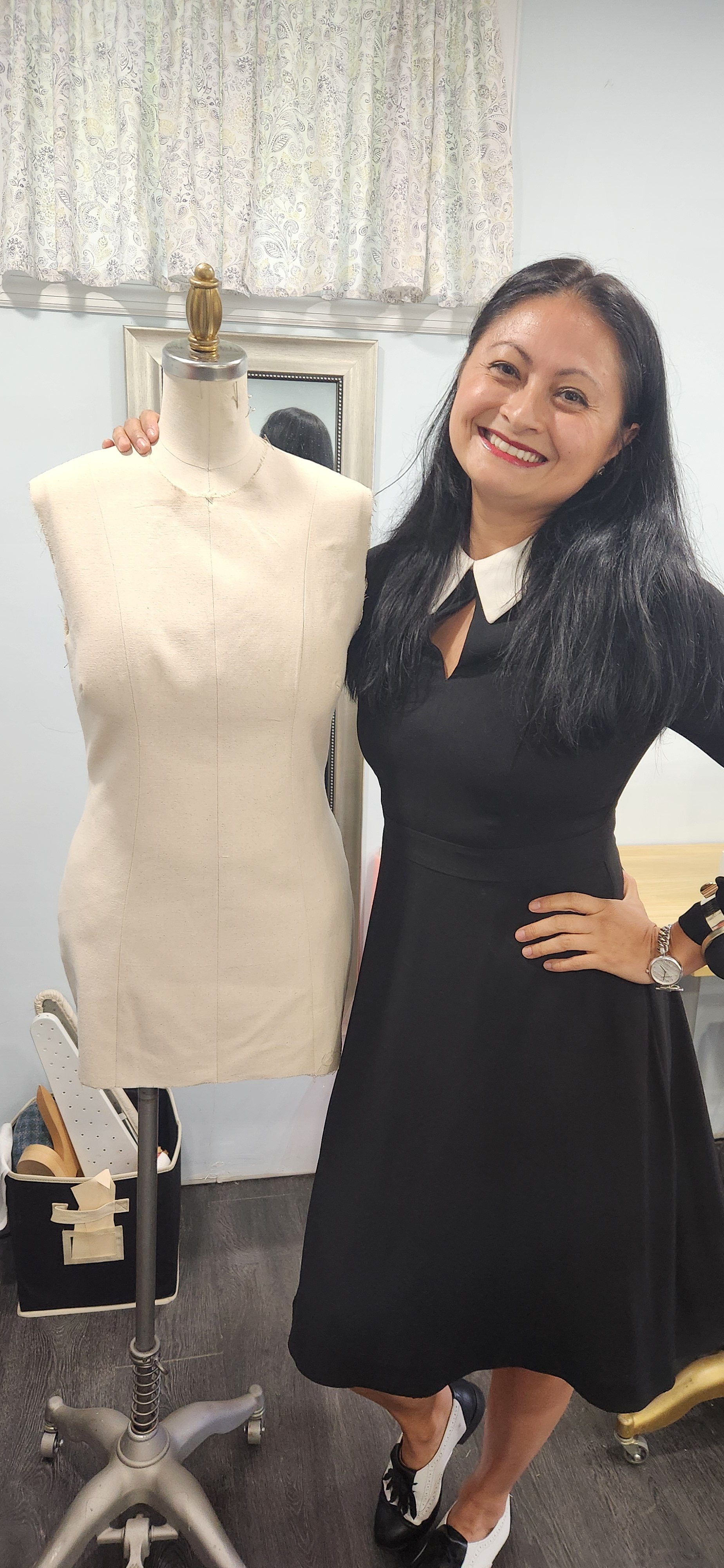 A smiling woman with long black hair standing next to a dress form in a room with light-colored walls and a window with patterned curtains. The woman is wearing a black dress with a white collar and black and white shoes. The dress form is beige and 