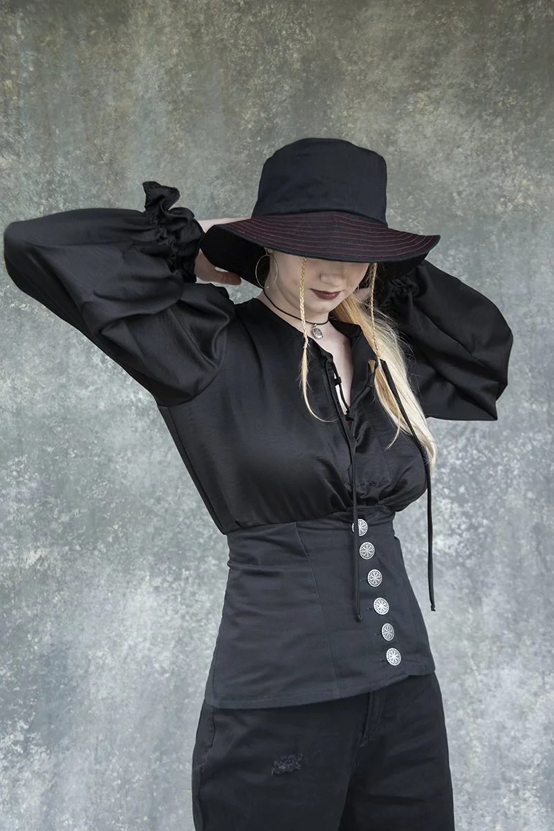 Woman in black outfit wearing a wide-brimmed hat with a red stripe, standing against a textured gray wall, with her hands behind her head.