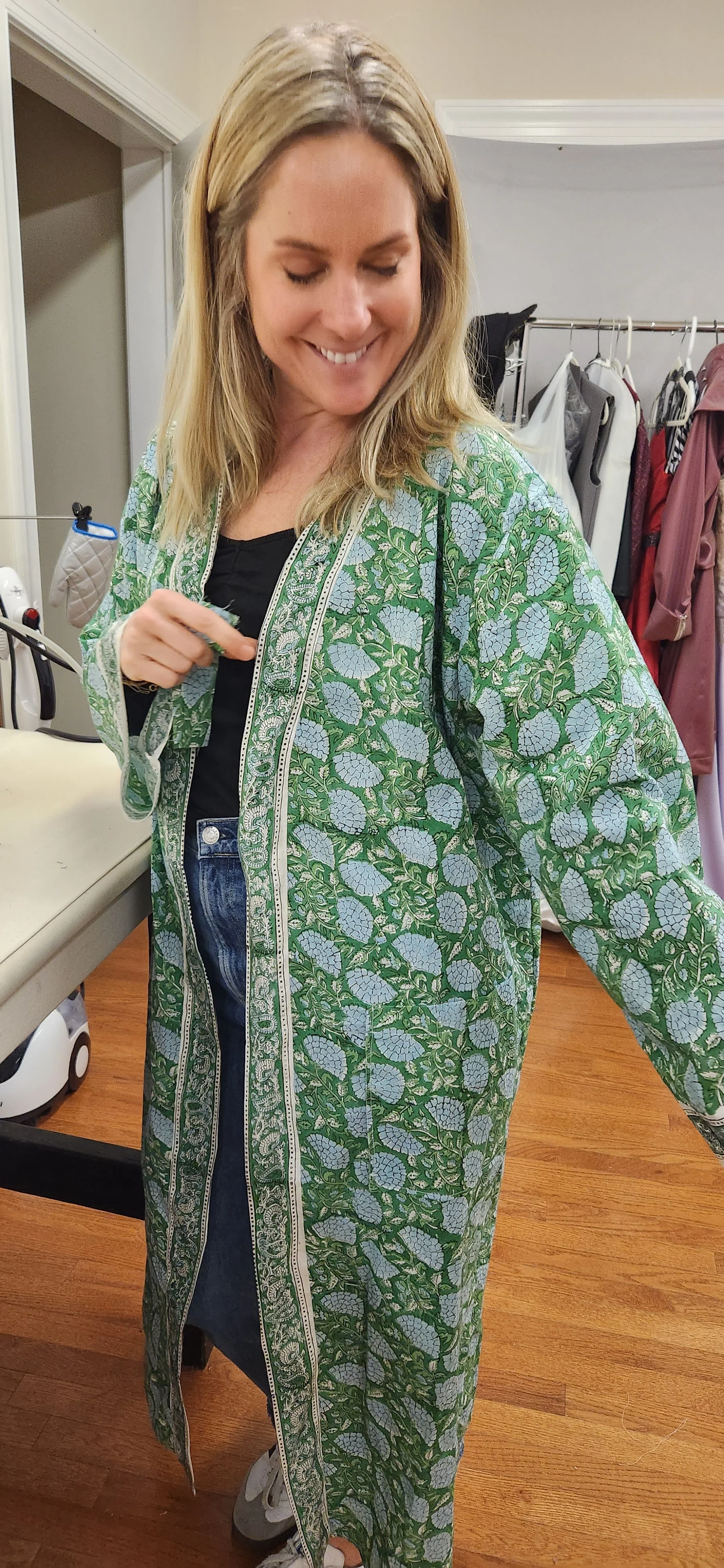 A woman smiling and wearing a long green and blue patterned kimono-style jacket, black shirt, jeans, and gray sneakers in a room with racks of clothing.
