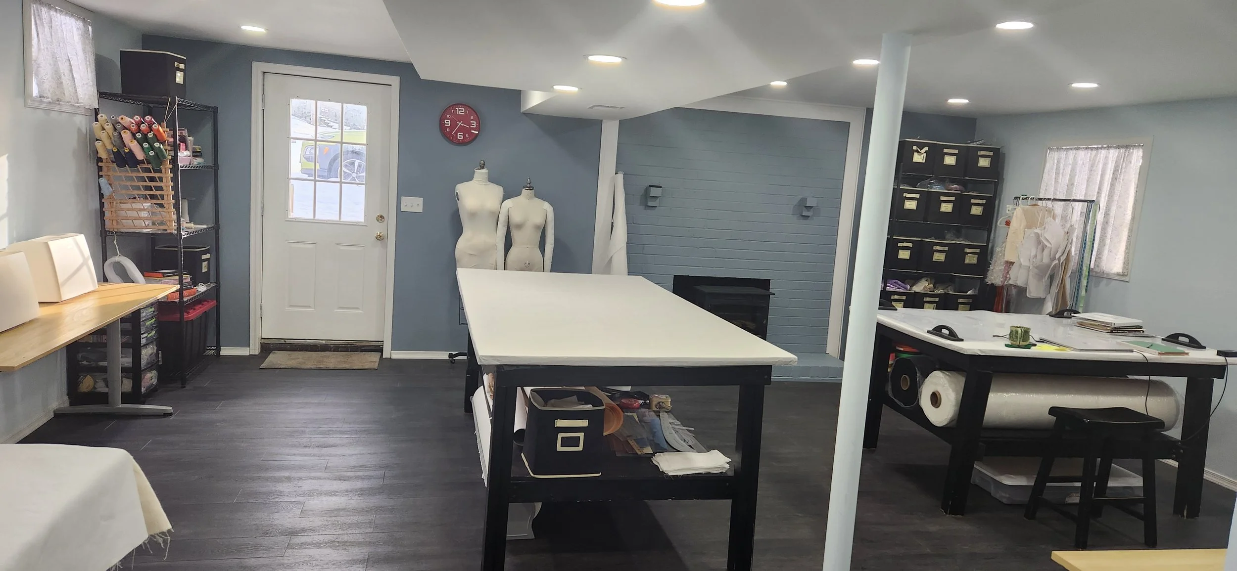Interior view of a sewing or craft workspace with tables, shelves filled with fabric and sewing supplies, dress forms, and a door with a window leading outside.