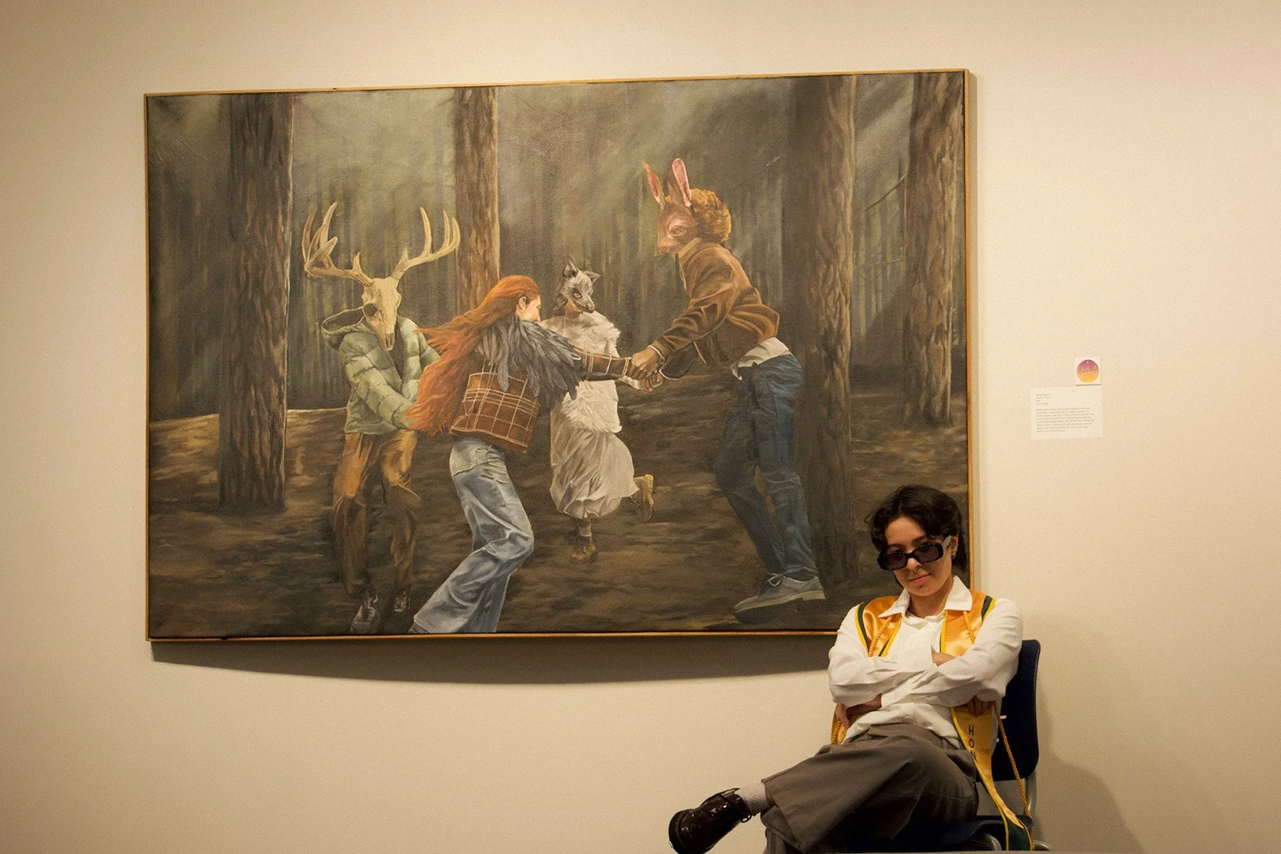 A person with glasses and a yellow stole sitting on a chair in front of a large forest scene painting with people dressed as animals holding hands and dancing in a forest.