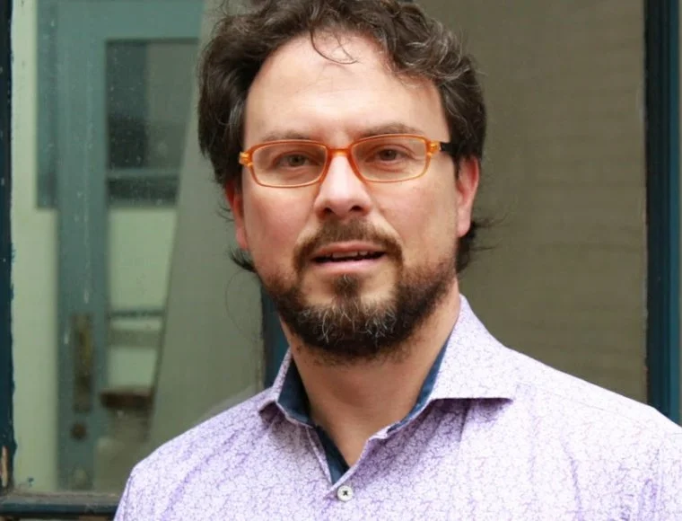 Hombre con barba, gafas naranja, camisa violeta y fondo con ventanas.