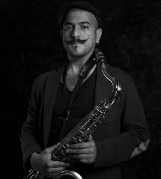 Hombre con bigote, barba y peinado peinado hacia un lado, sosteniendo un saxofón, con fondo oscuro.