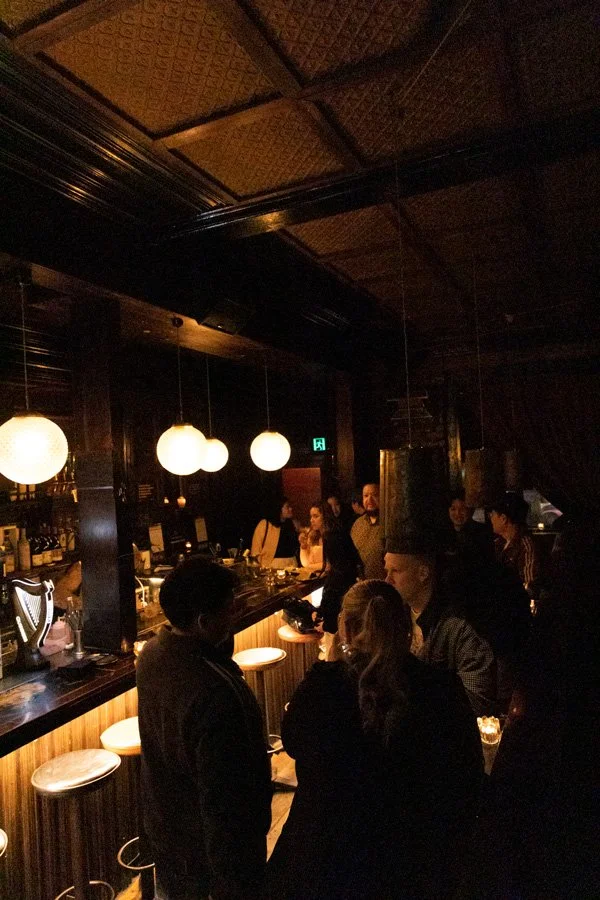 Dimly lit bar with hanging spherical lights, people socializing at the counter and standing around, dark wood decor.