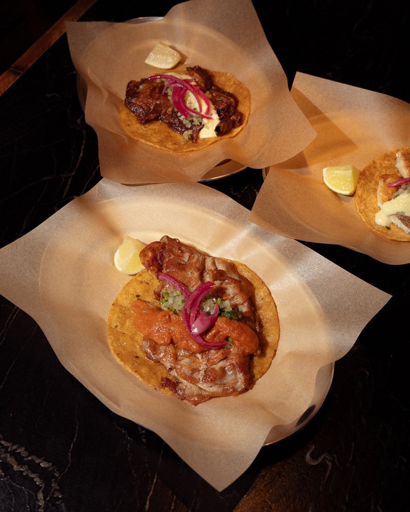 Three tacos on parchment paper with lemon wedges, showing meat, pickled red onions, and garnish.