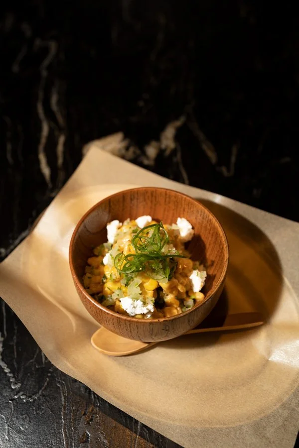 A wooden bowl of corn salad garnished with green onion slices, served on parchment paper with a small wooden spoon.