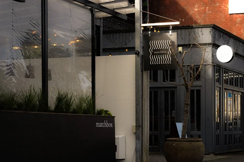 Exterior view of a cafe or restaurant called 'matchbox' with a black facade, a leafless tree in a large pot, and large glass windows reflecting the street.