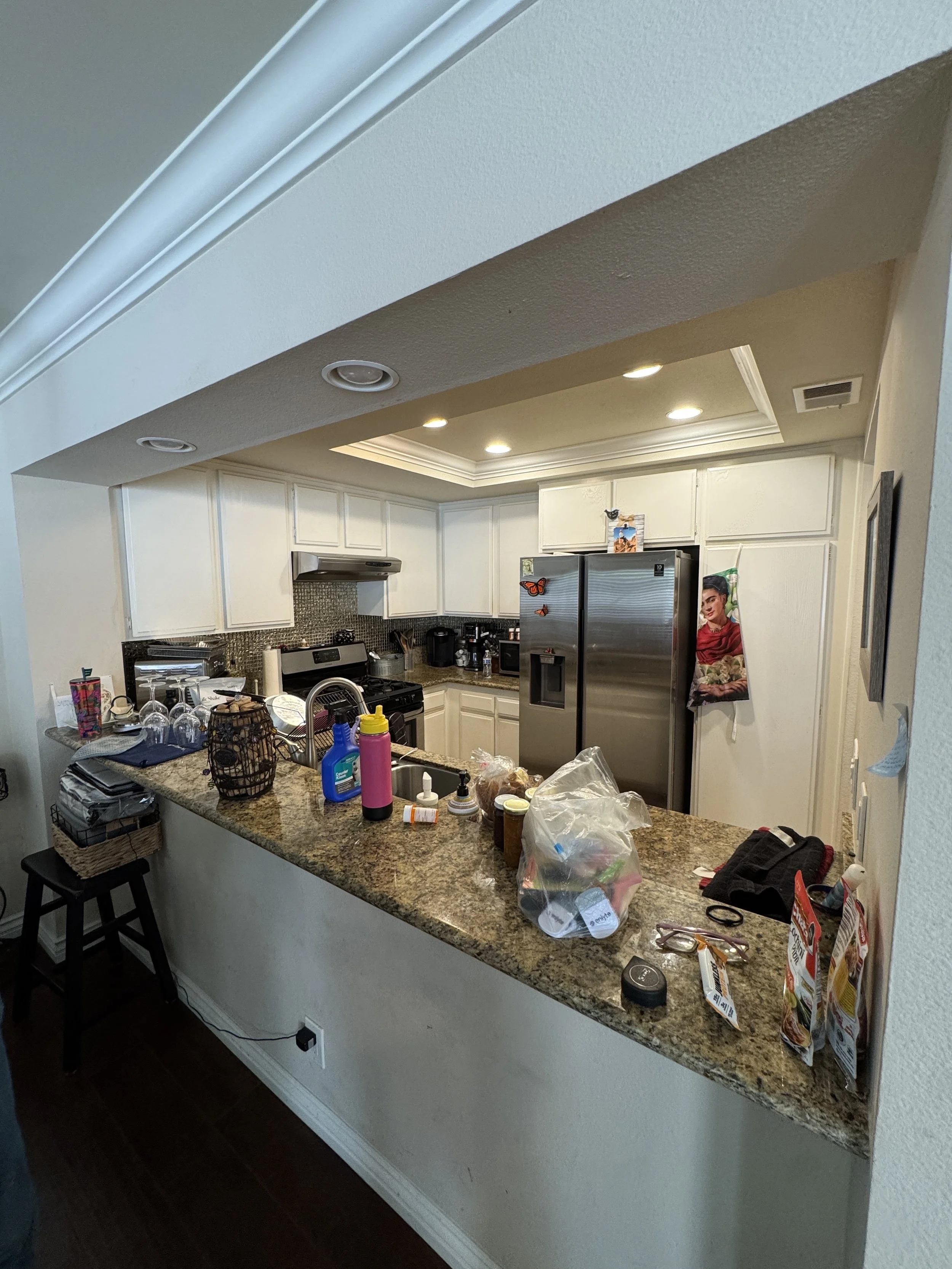 Kitchen with granite island counter cluttered with bottles, paper towels, food packages, and kitchen utensils, white cabinets, stainless steel refrigerator, black stove, and dim recessed lighting.