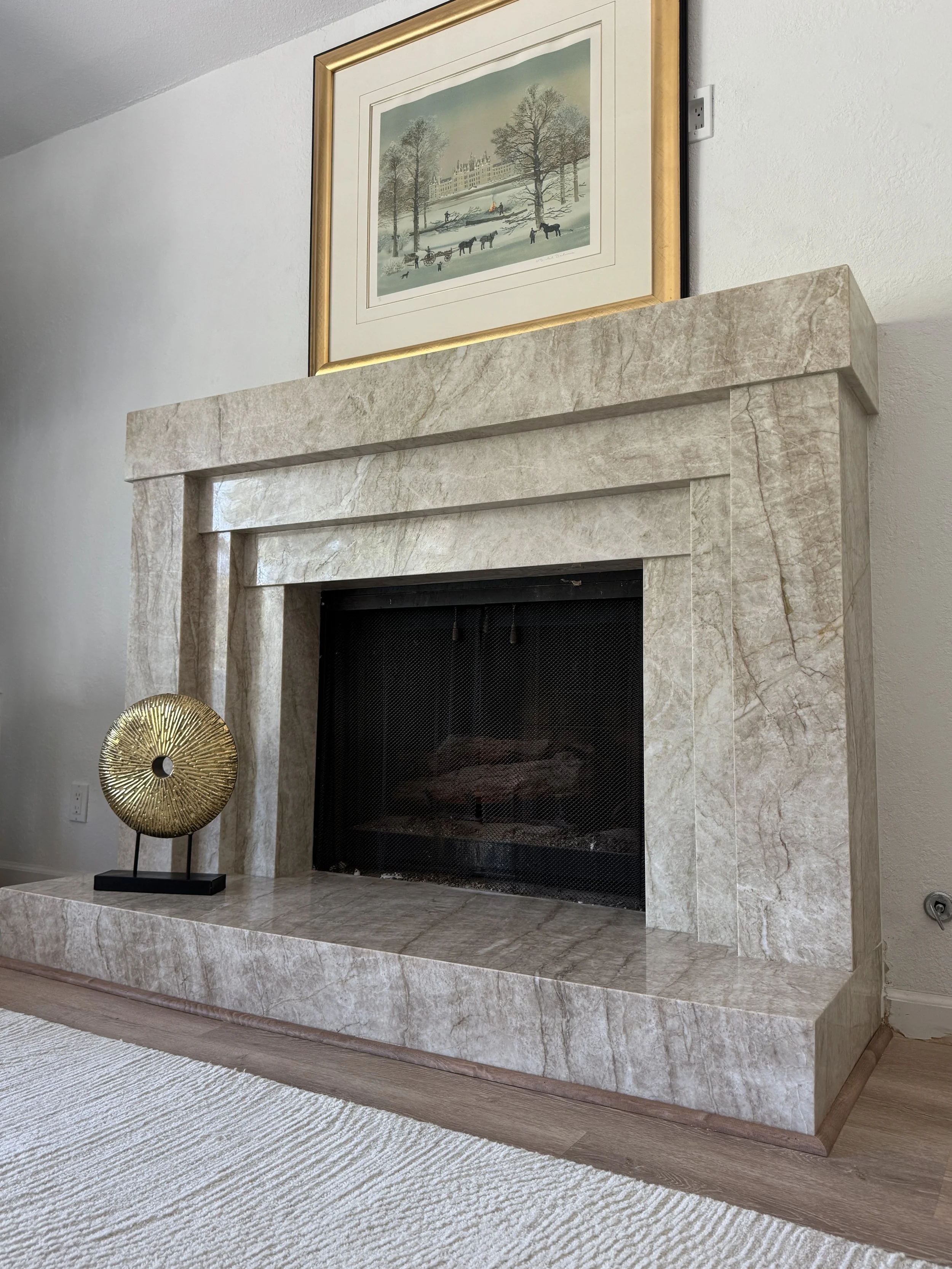 A beige marble fireplace with a rectangular design, set against a plain white wall. The fireplace has a black metal screen and is surrounded by a matching marble mantel and hearth. The floor is light-colored wood.