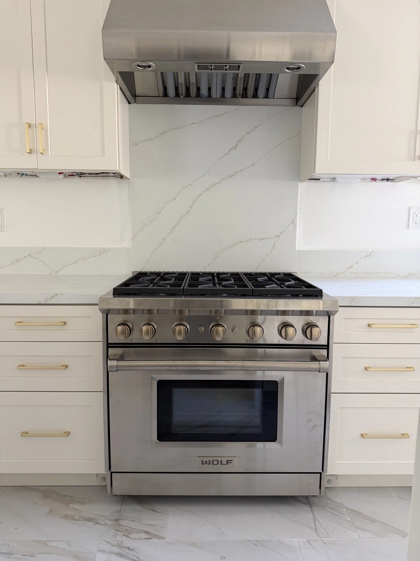 Stainless steel Wolf gas range with marble countertop and white cabinetry with gold handles in a modern kitchen.