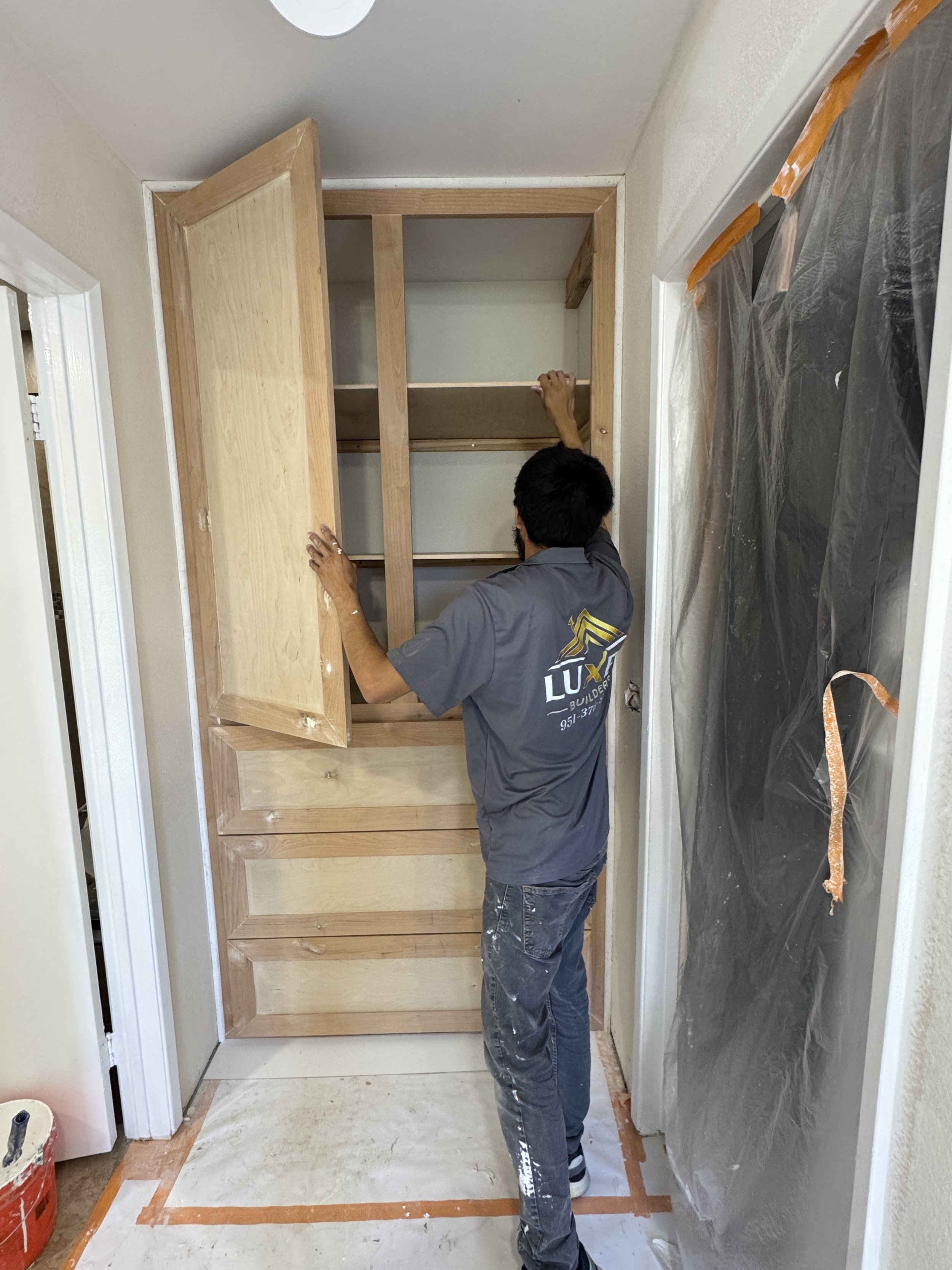 A worker installing a wooden cabinet in a room under construction or renovation. The worker is facing the cabinet, which is being attached to the wall. Protective plastic covers parts of the wall on the right side, and the floor is covered with a protective sheet with orange tape outlining the area.