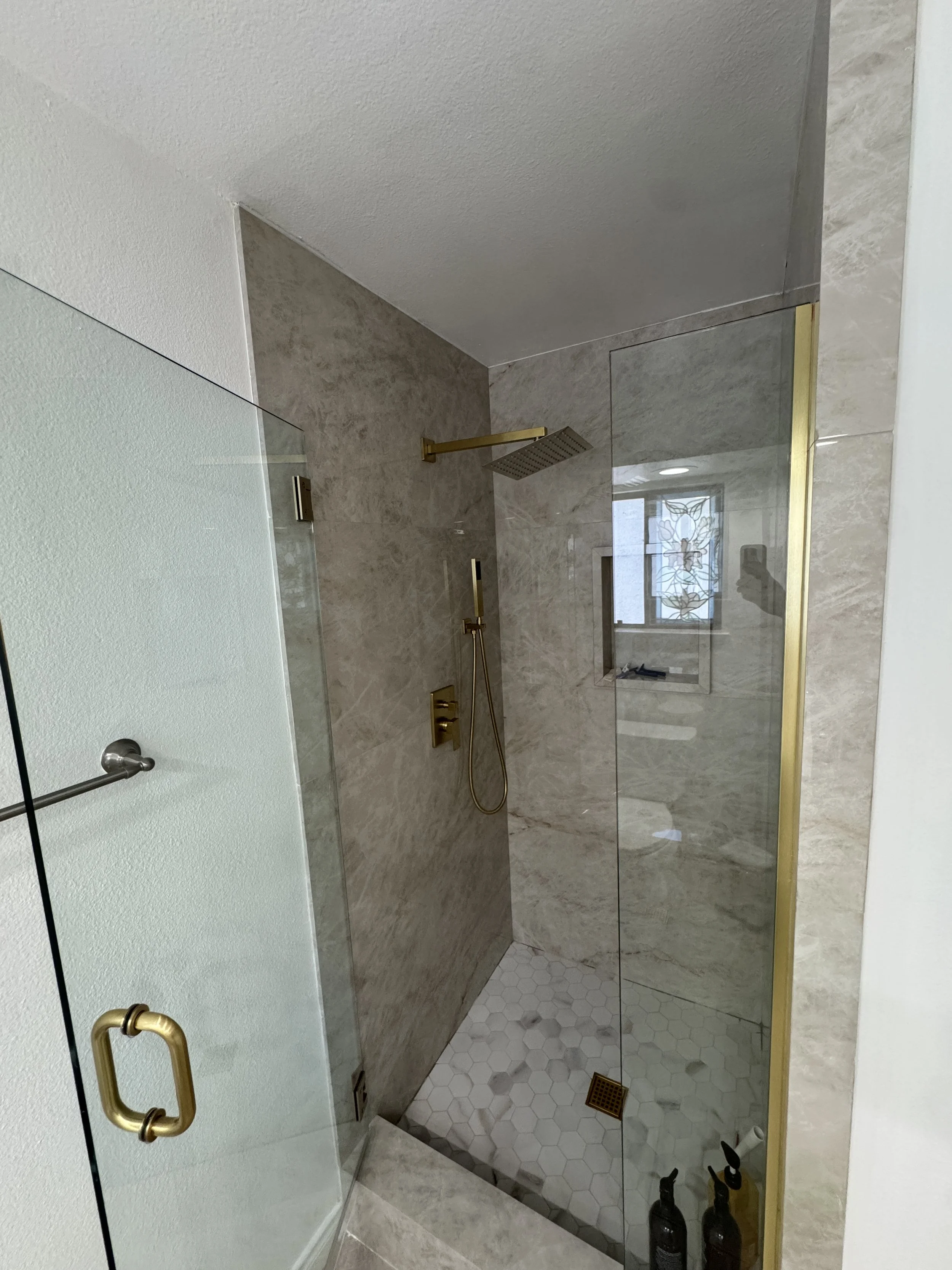 Bathroom shower with a brass showerhead and handheld. Marble walls, glass door with gold handle, hexagon and square tile flooring, window with stained glass, and bottles of shower products on the floor.