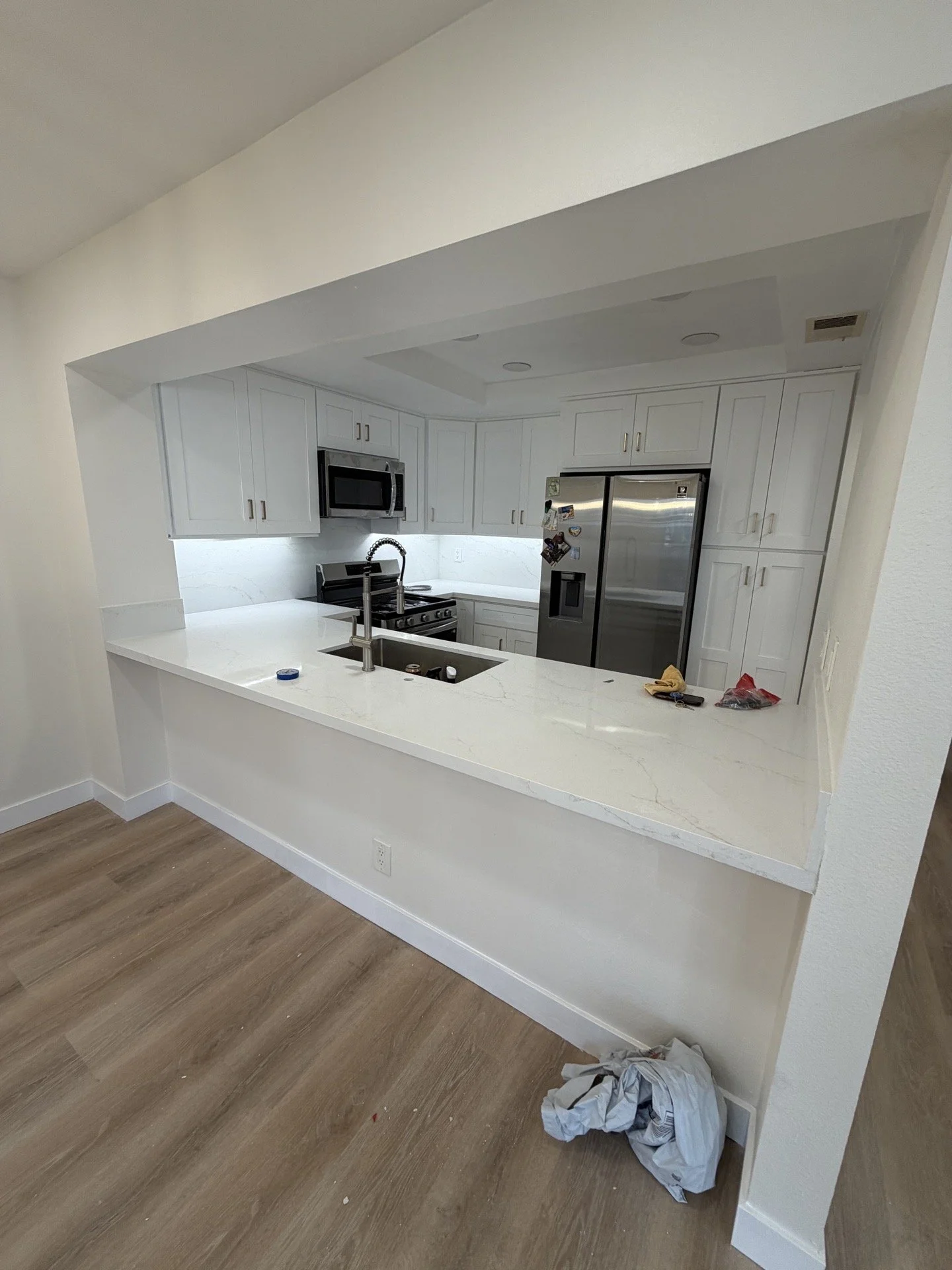 Modern kitchen with white cabinets, stainless steel appliances, and a white countertop with a sink. There are some tools and items on the counter, and a pile of laundry or cloth on the floor.