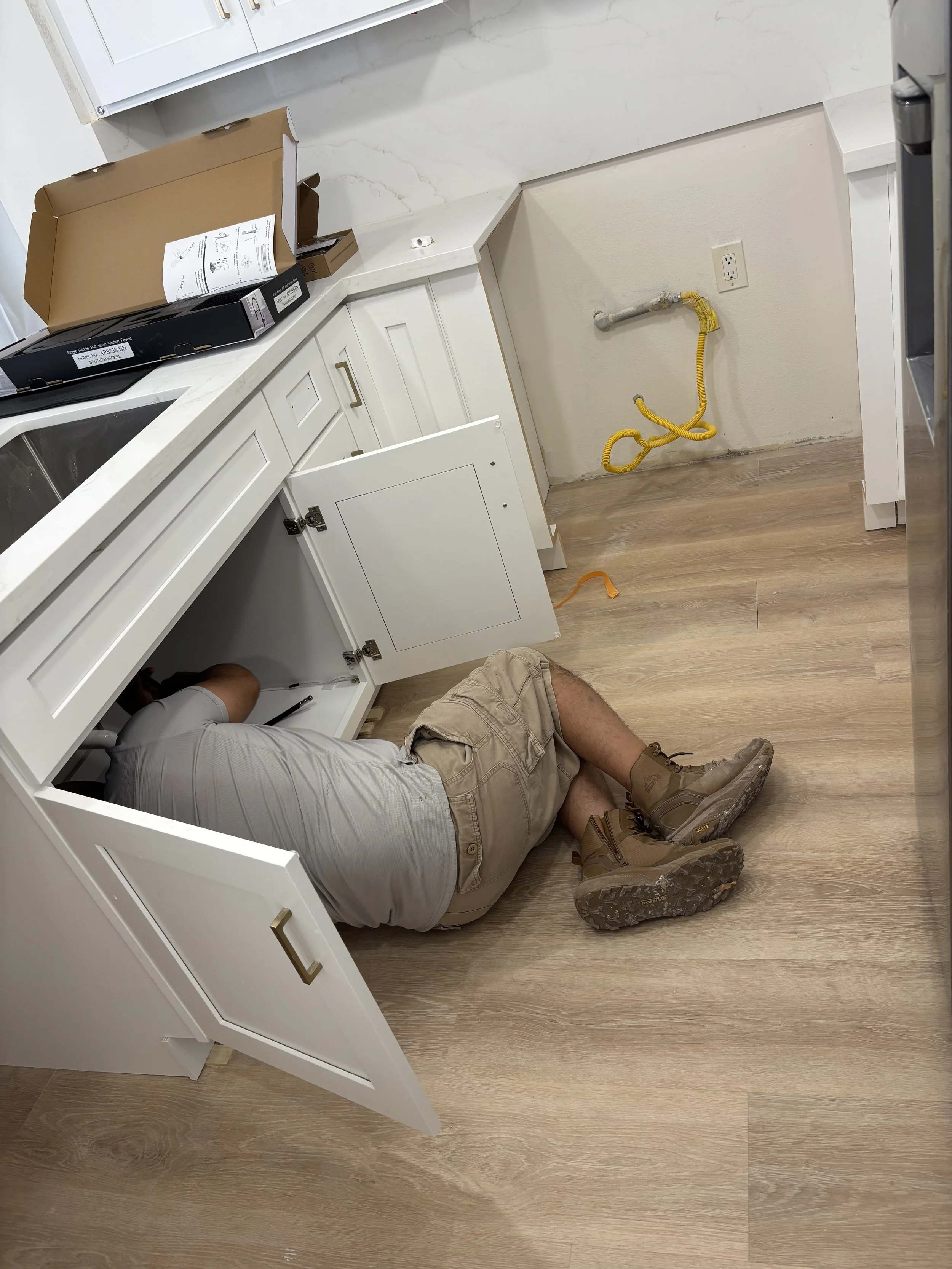 Person kneeling on the floor working on kitchen cabinets, with open cabinet door, and cardboard box on the countertop nearby. The wall has an electrical outlet and a pipe with yellow flexible tubing.