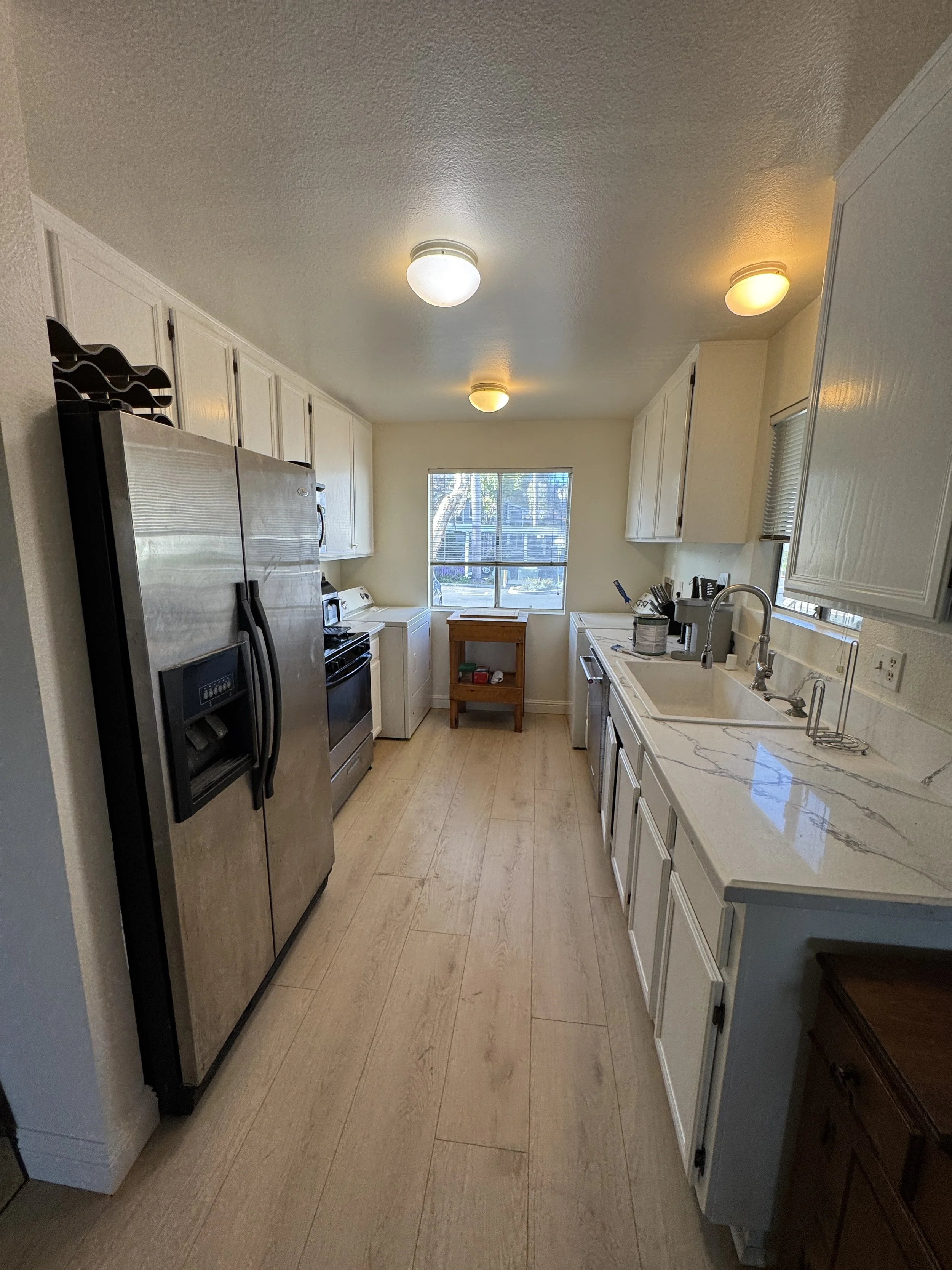 A kitchen with white cabinets, a stainless steel refrigerator, a stove, a sink with a window above it, and beige tile flooring. There are ceiling lights on the textured ceiling and some kitchen utensils and a small wooden table by the window.