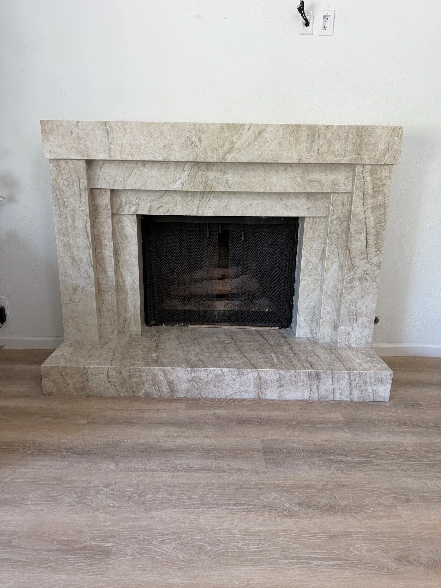 A beige marble fireplace with a rectangular design, set against a plain white wall. The fireplace has a black metal screen and is surrounded by a matching marble mantel and hearth. The floor is light-colored wood.