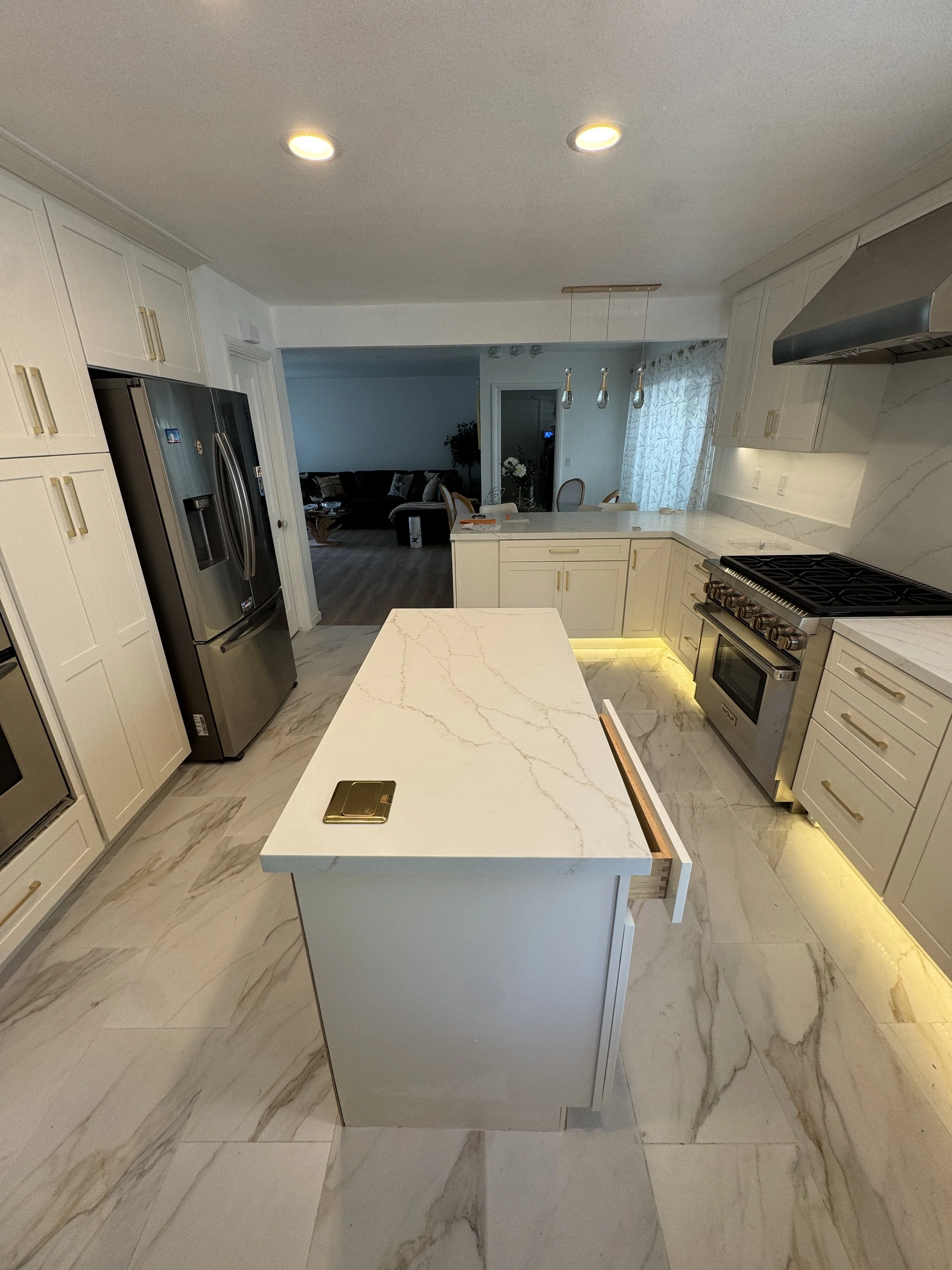 Modern kitchen with white marble countertops and flooring, stainless steel appliances, and white cabinets with gold handles. There is a kitchen island in the foreground and a dining area with a chandelier and door to a living room with a dark sofa.