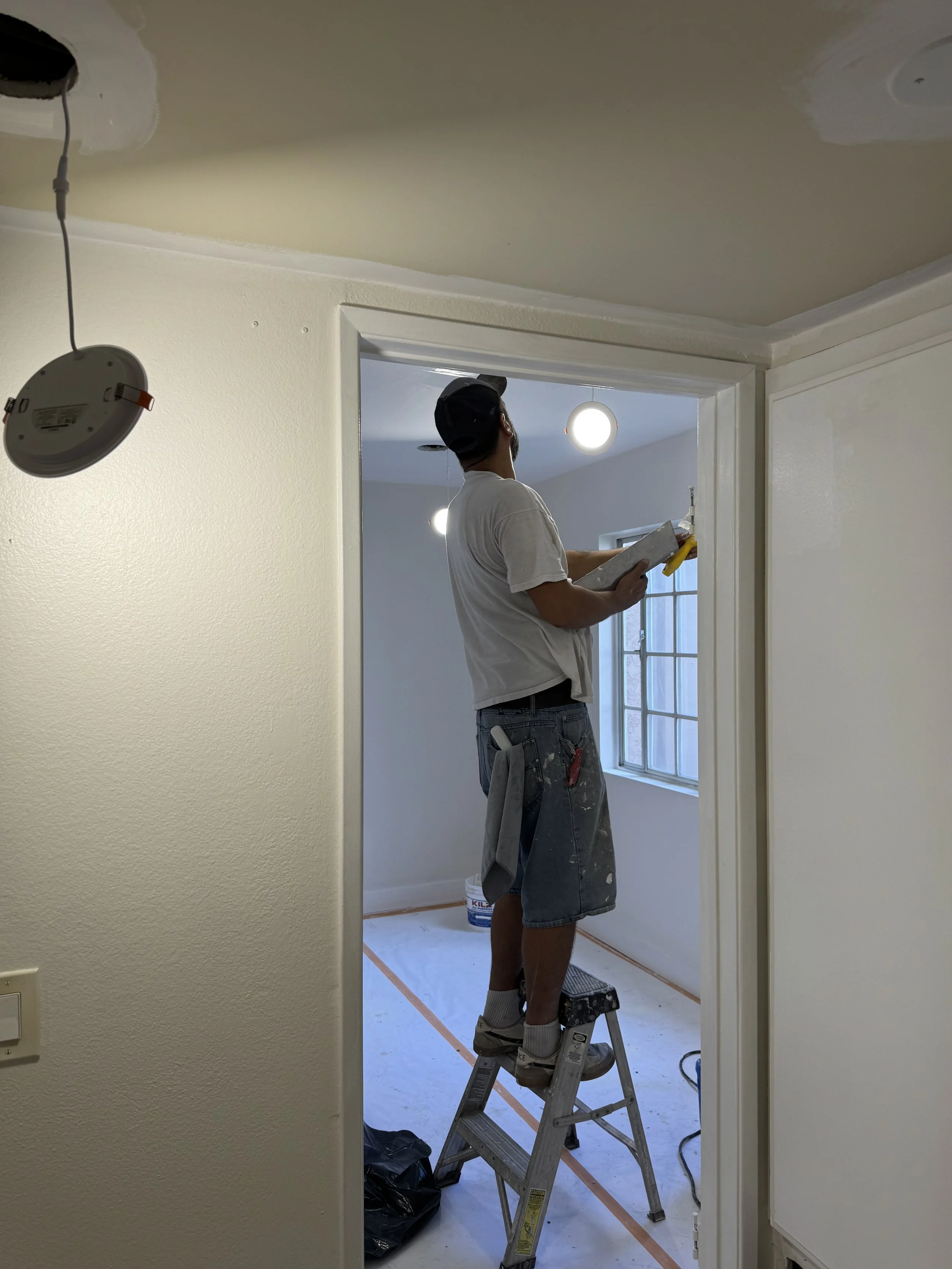 A man standing on a step ladder, painting a doorway frame during a home renovation. He is wearing a white t-shirt, shorts, and a cap, and is holding a paintbrush and a paint tray. There are paint cans on the floor and plastic sheeting covering the floor.