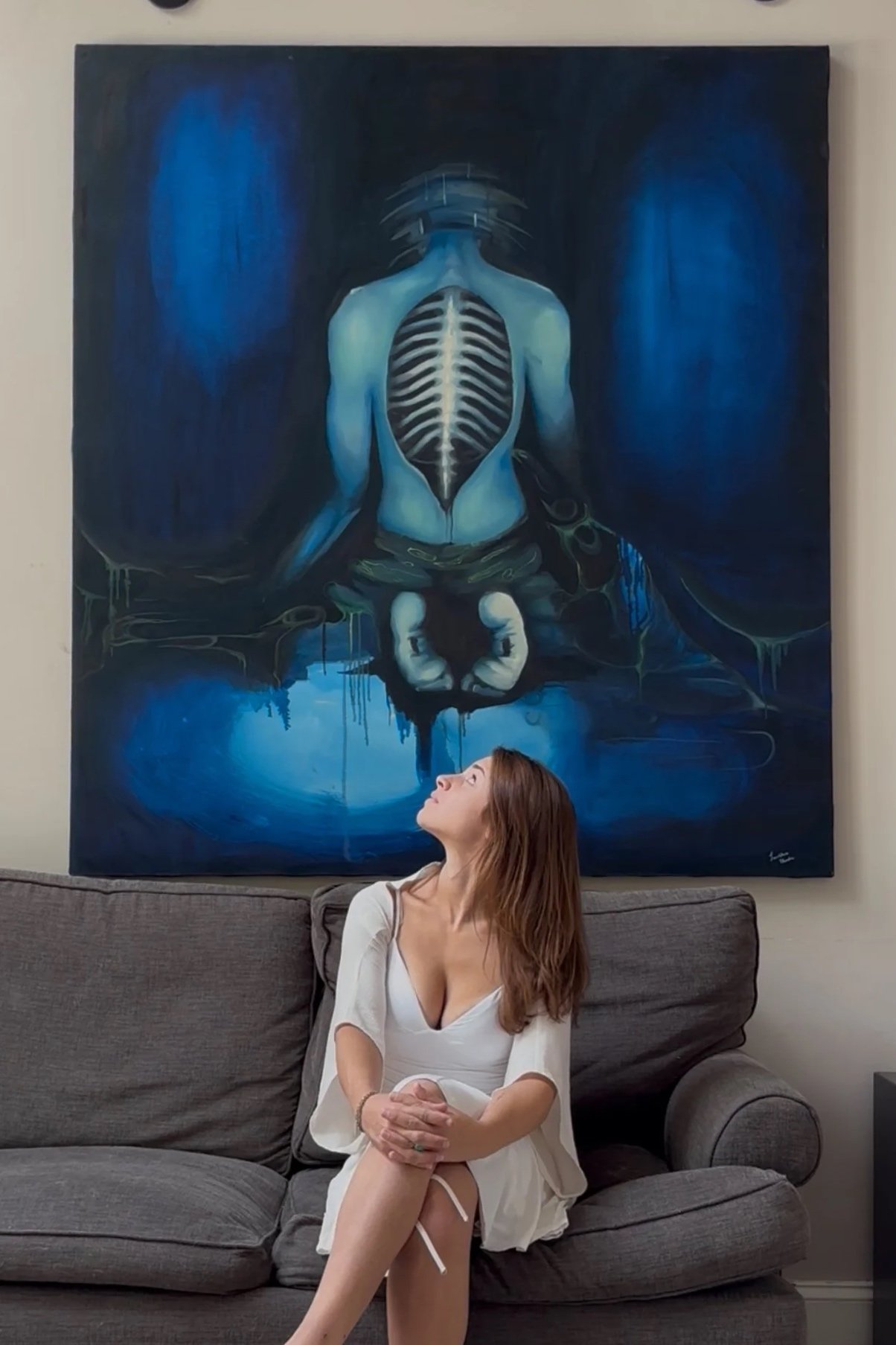 A woman sitting on a gray couch looking up at a large painting behind her. The painting features a blue-toned surreal image of a person with an exposed rib cage and a skeleton of hands in a mirror reflection, with dark and glowing blue hues.