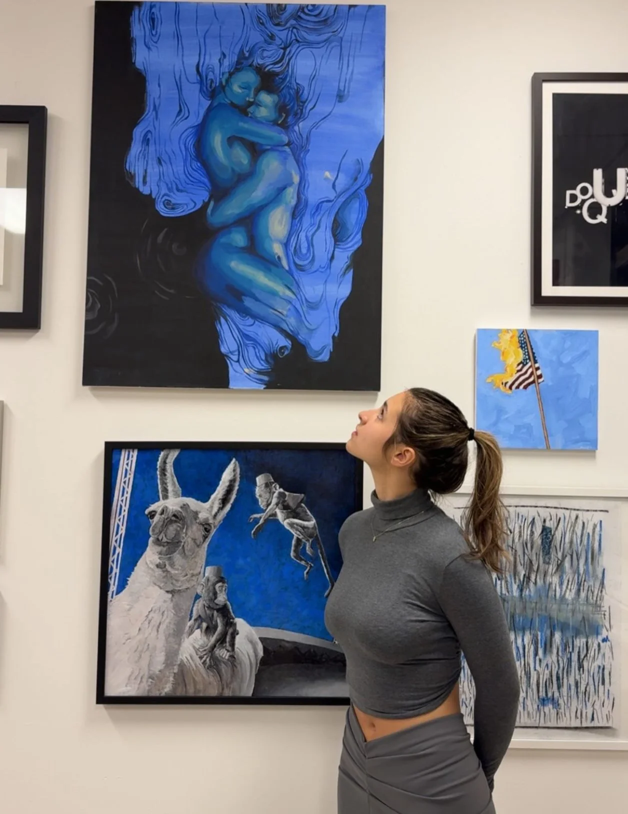 A woman with a ponytail looking at paintings on a gallery wall, including a blue-toned artwork of two people embracing, a drawing of a llama with other animals, and a small American flag painting.
