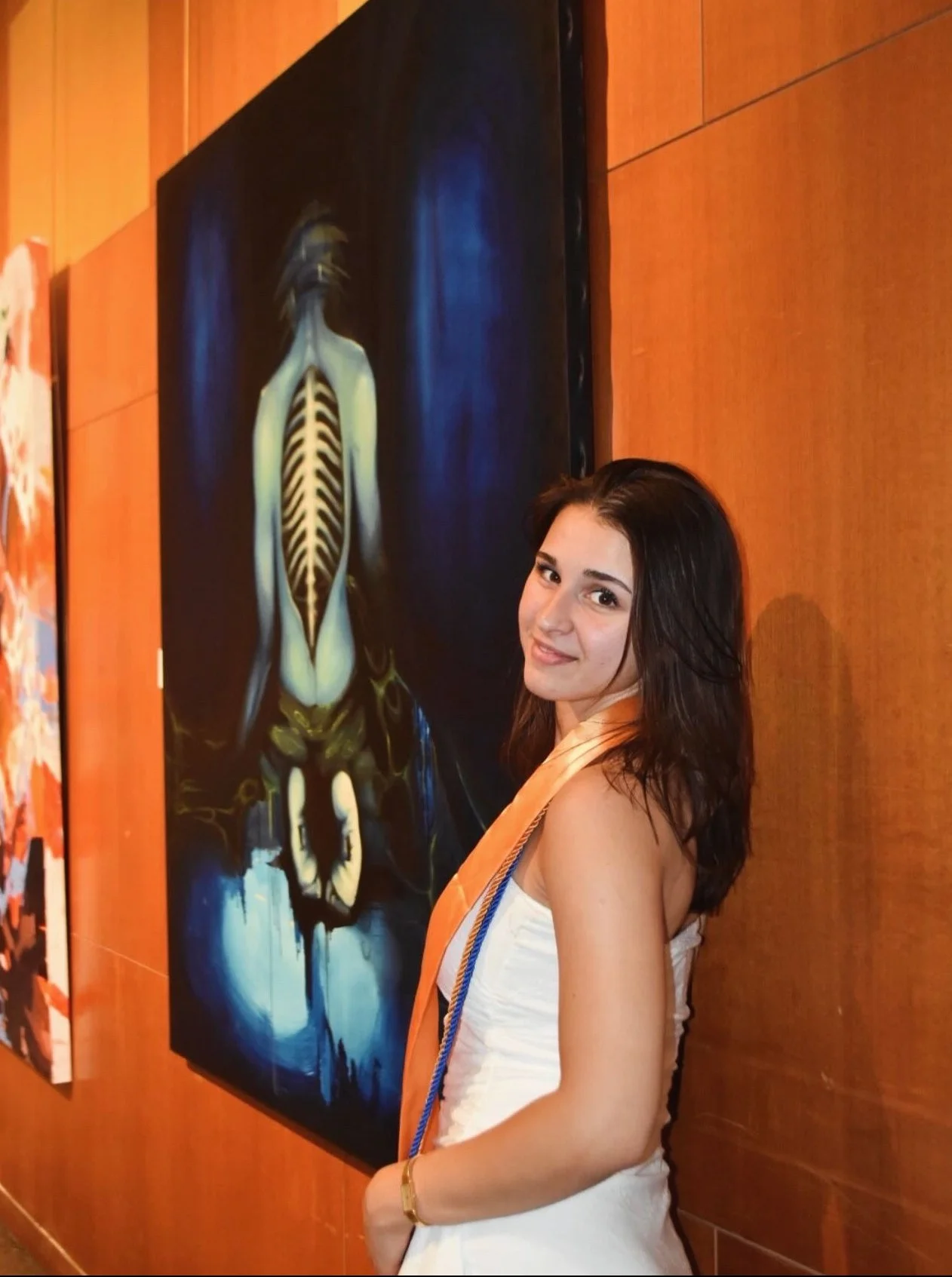 Young woman with long dark hair wearing a white dress and a peach-colored sash, standing next to a large, dark-themed painting of a skeletal figure with a glowing blue background at an art gallery.
