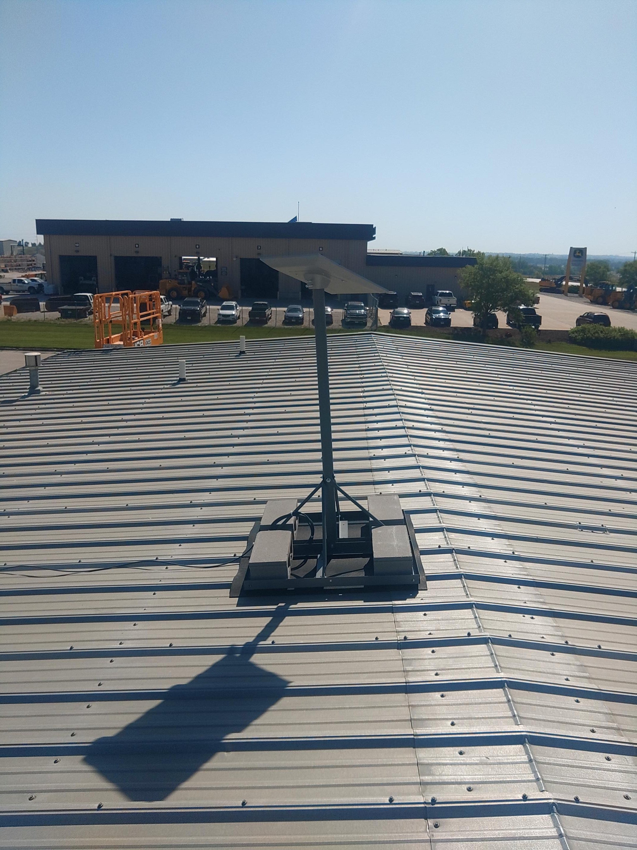 View of a rooftop with a mounted antenna or satellite dish in the foreground, a parking lot with cars and construction equipment, a building with a garage, a large sign with a logo and a few trees, under a clear blue sky.