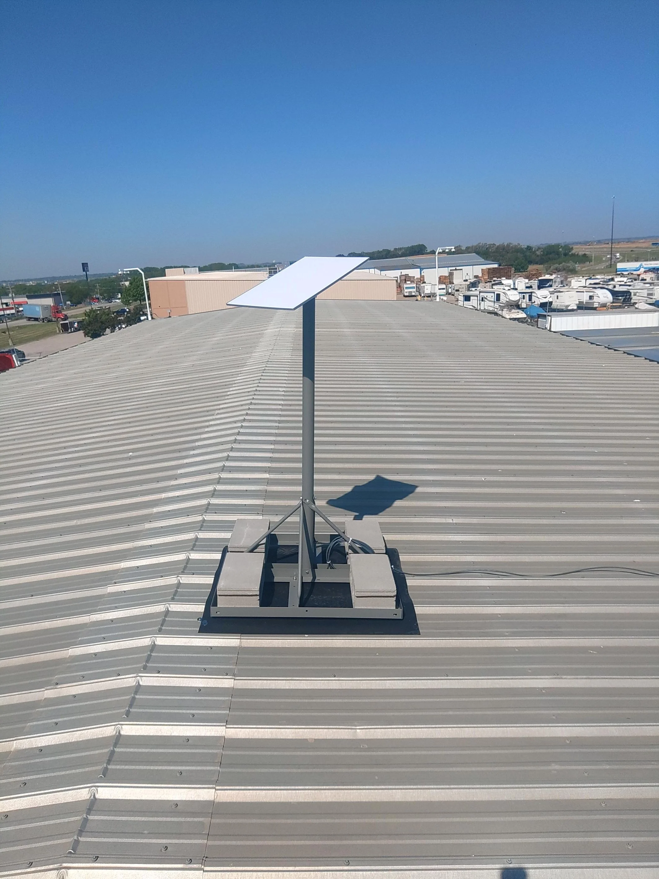 A small solar panel mounted on a metal pole on a flat, metal roof with a scenic background of trees, buildings, and a clear blue sky.