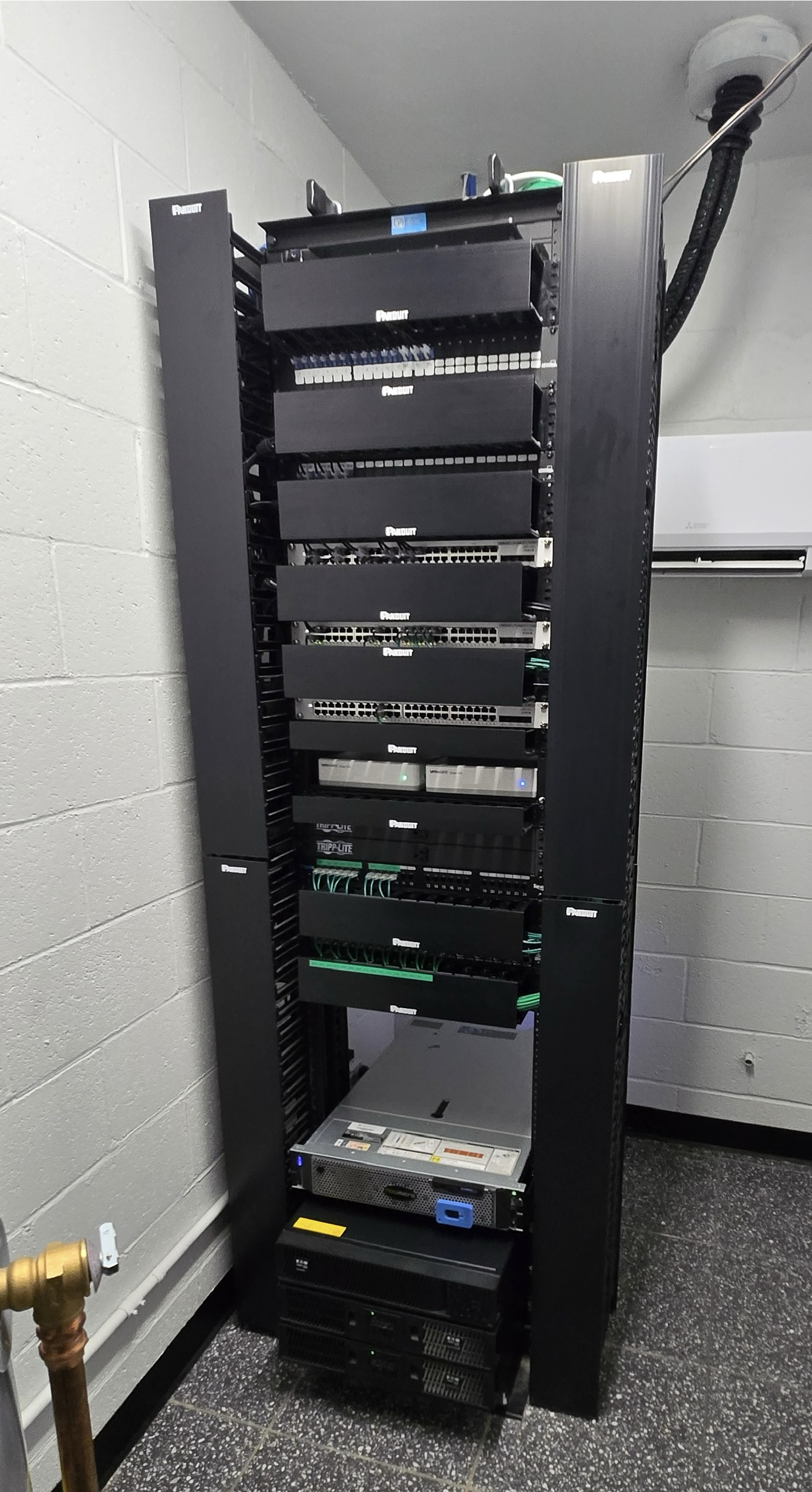 A tall black server rack containing multiple network and server equipment installed in a data center or server room, with cables and devices visible at the bottom.