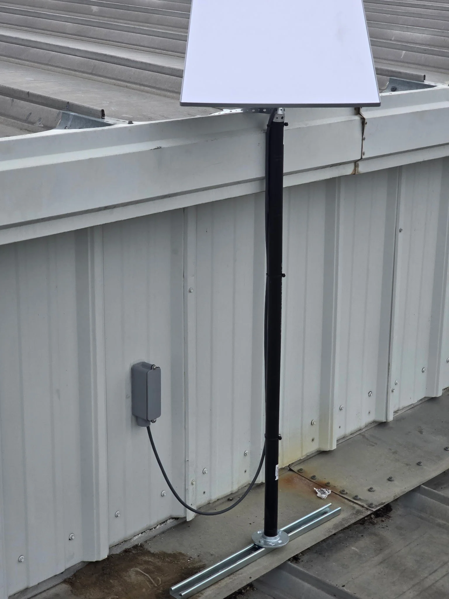 A white electronic device mounted on a metal rail against a metal building wall, connected by a black pole and cable.