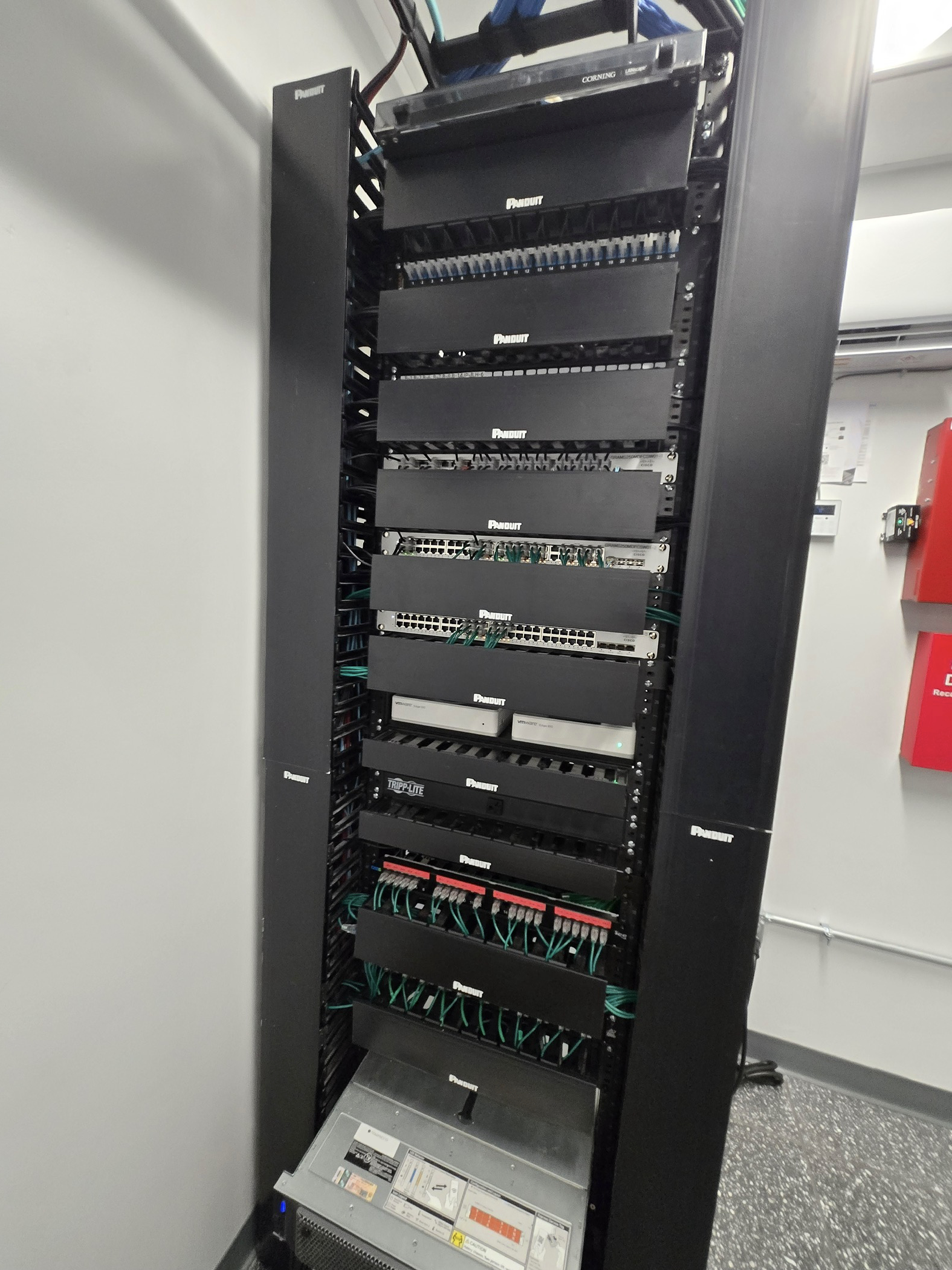 A tall black server rack with multiple networking and computer hardware components, including switches, routers, and power supplies, situated in a room with a gray wall, a window, and a speckled black floor.