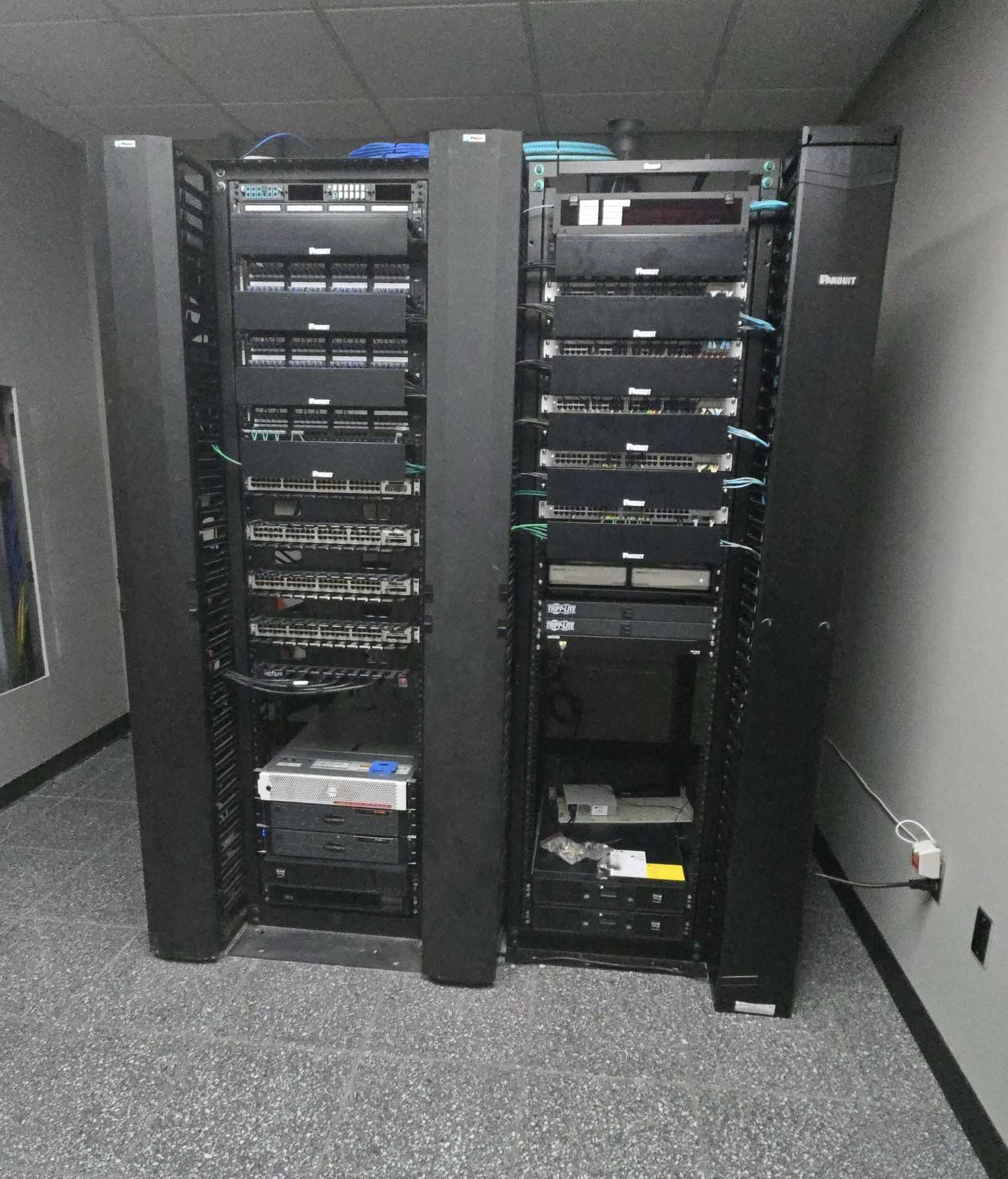 A server rack in a data closet with multiple black electronic equipment units, network cables, and power cords.