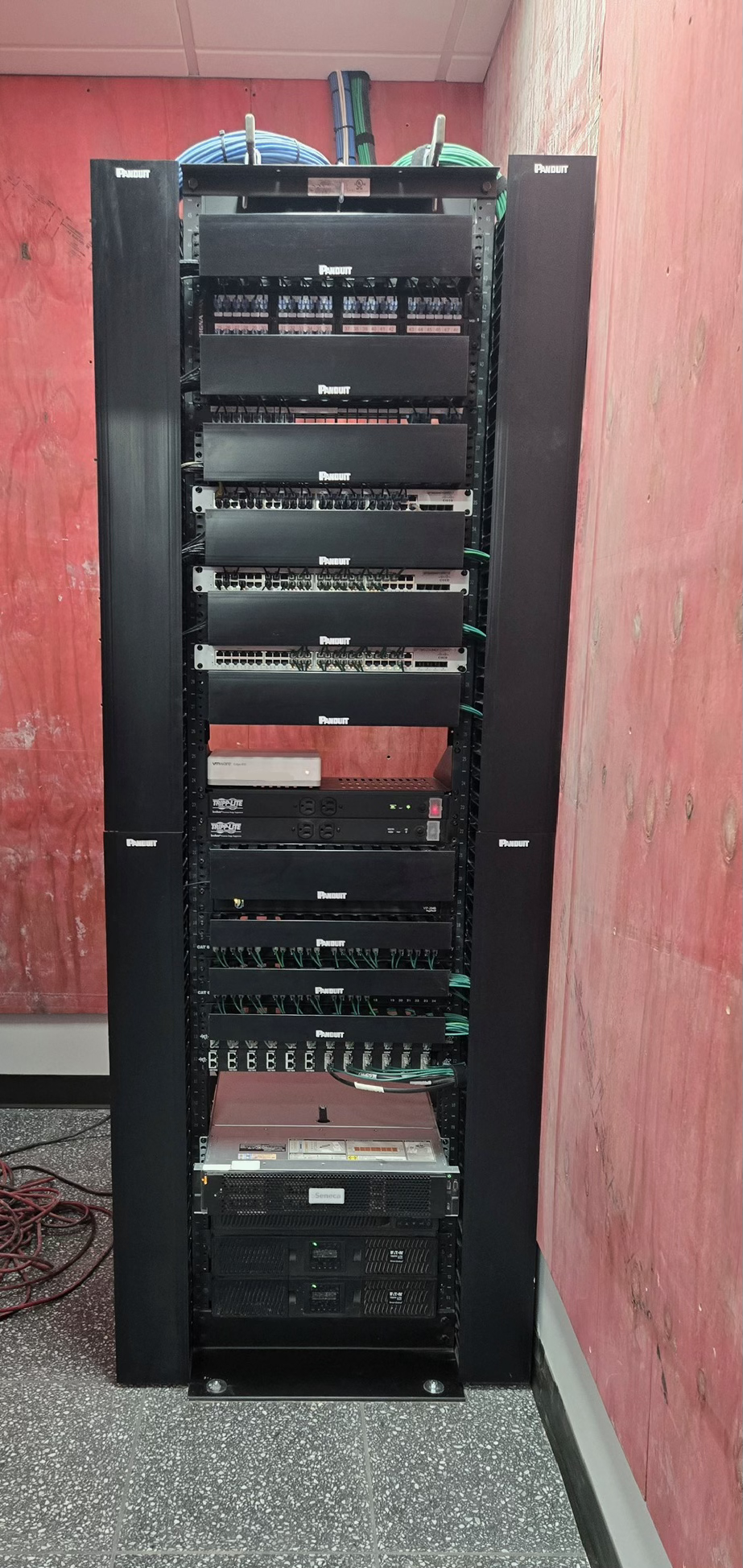 A server rack with networking and power equipment, including patch panels, switches, and power supplies, located in a room with pinkish-red walls and speckled flooring.