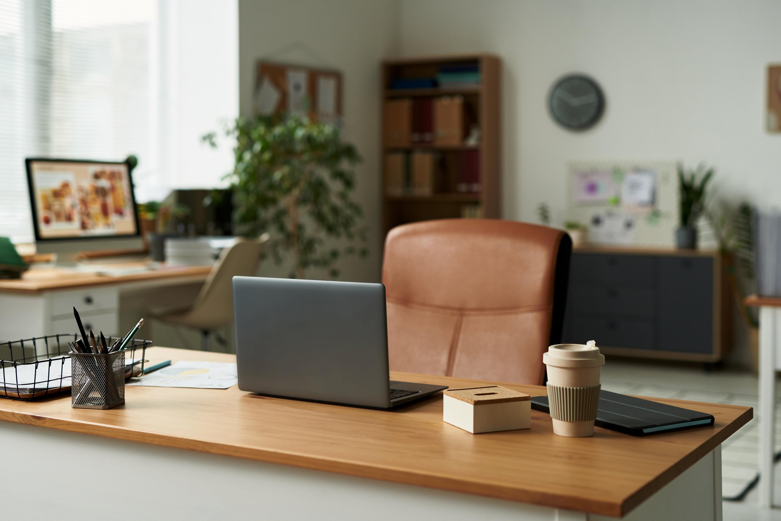 Office workspace with a wooden desk, an open laptop, a coffee cup, a wire pen holder, and a notebook. In the background, there are shelves, a clock on the wall, and plants by the window.