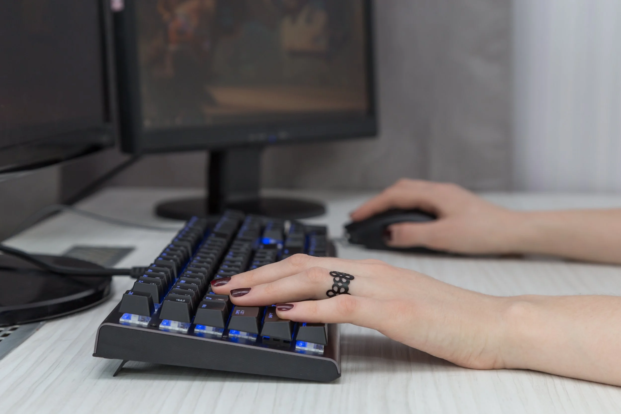 Person with painted nails and a ring on their finger typing on a backlit mechanical keyboard at a desk with a computer monitor in the background.