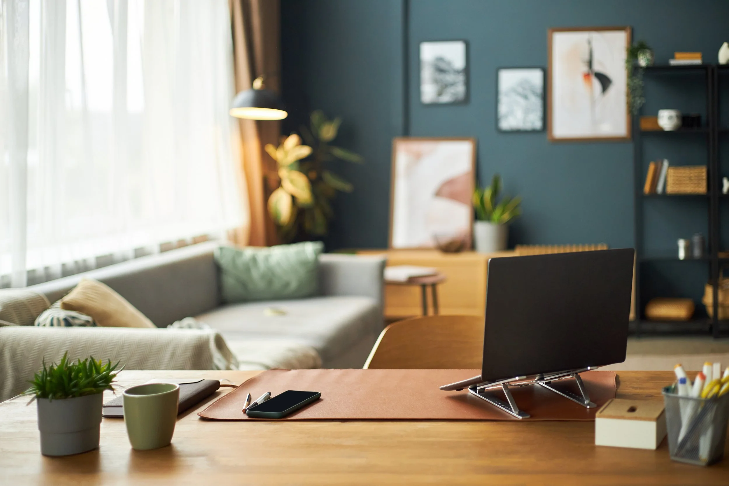 A workspace with a wooden desk holding two cups, a smartphone, pens, and a potted plant, with a backdrop of a living room featuring a sofa, cushions, a lamp, and gallery wall.